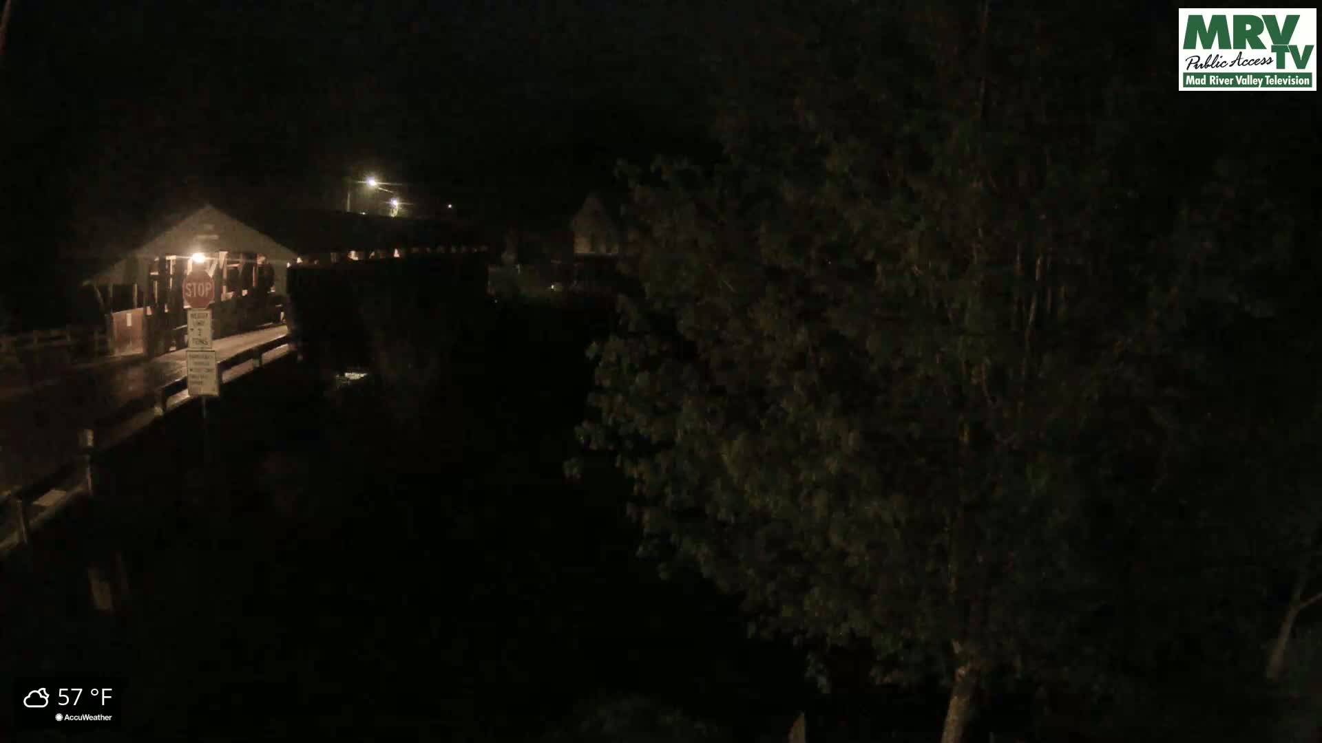 A nighttime view of a covered bridge and surrounding trees under a dark sky, with a temperature of 57°F.