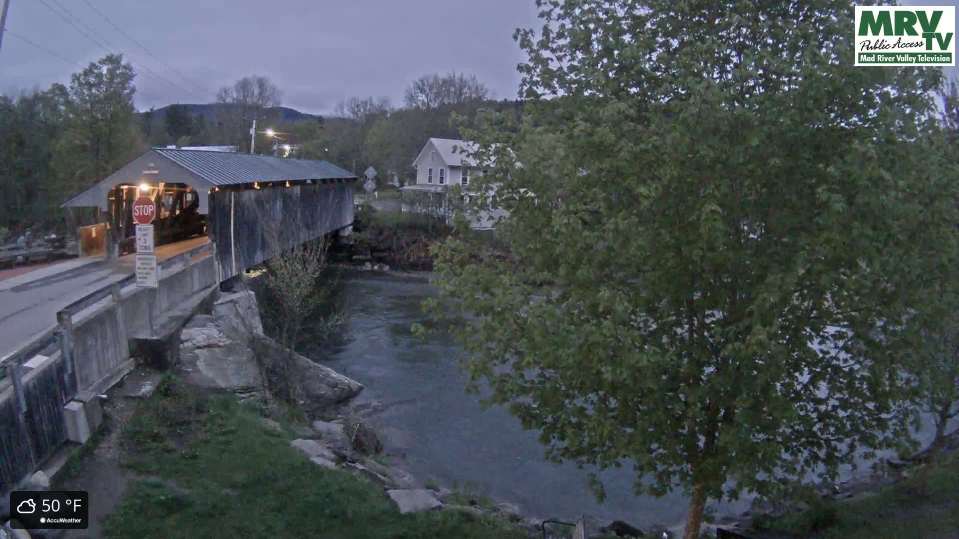 A covered bridge spans a river, with a house visible in the background under a cool, 50°F sky.