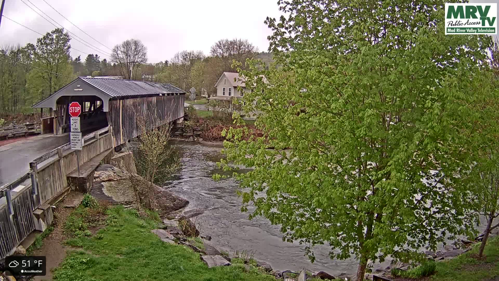 A covered bridge spans a flowing river under an overcast sky, with lush green trees and a house visible in the background.