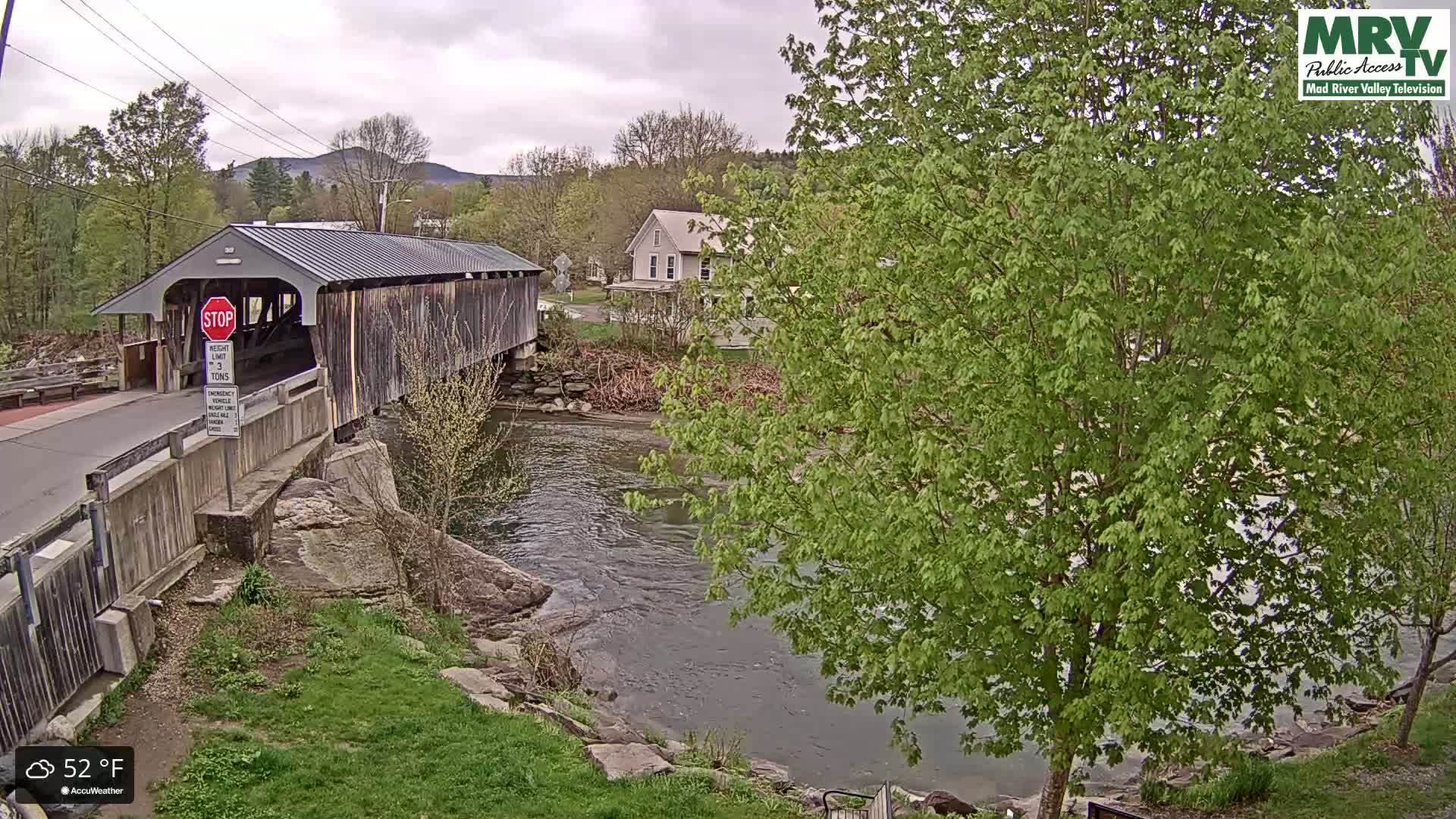 A covered bridge spans a river under an overcast sky, with lush green foliage surrounding the waterway and a building visible in the background.