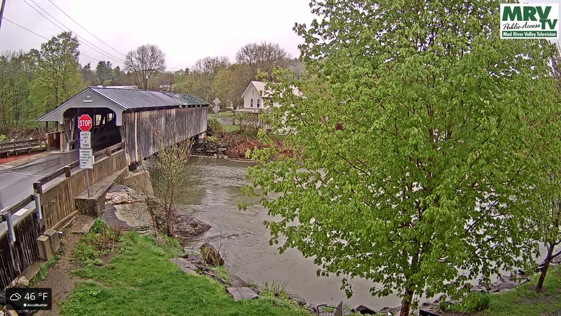 A covered bridge spans a muddy river under an overcast sky, with lush green trees and a house visible in the background.
