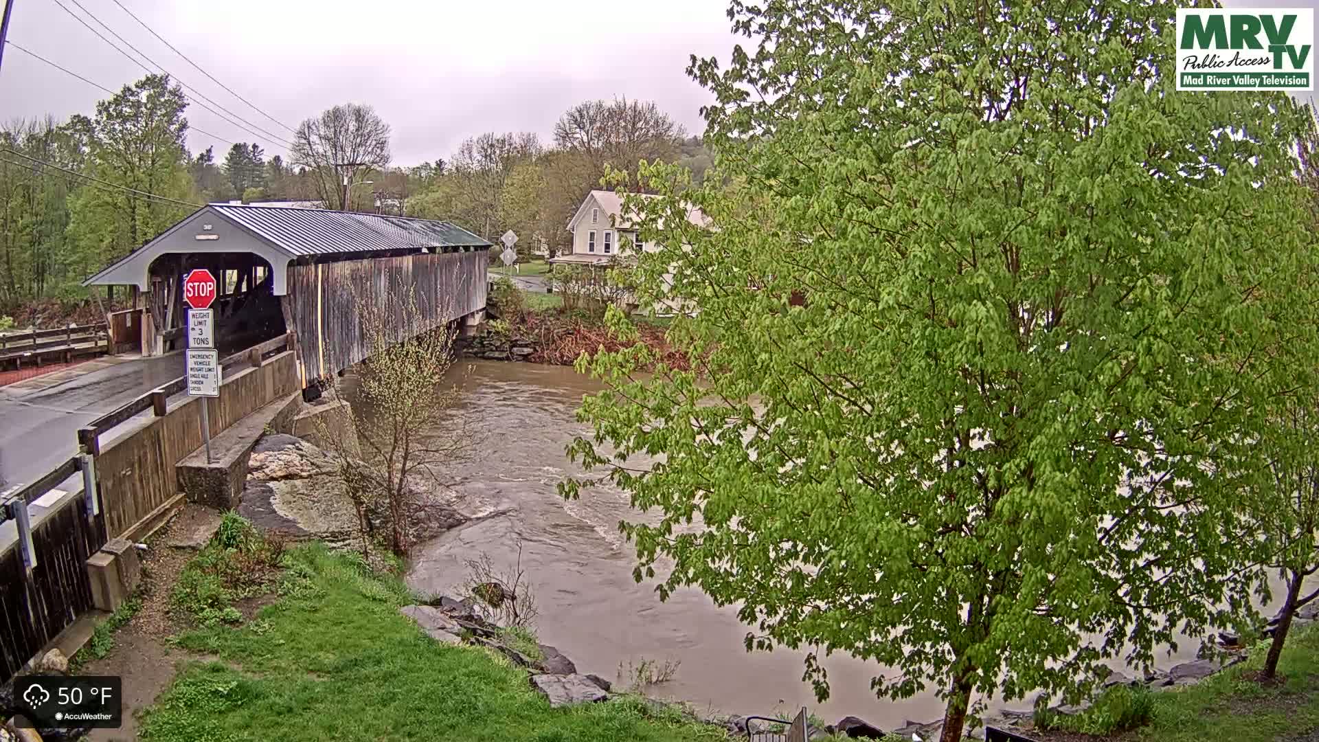 A covered wooden bridge spans a muddy river under an overcast sky, with lush green trees lining the riverbank.