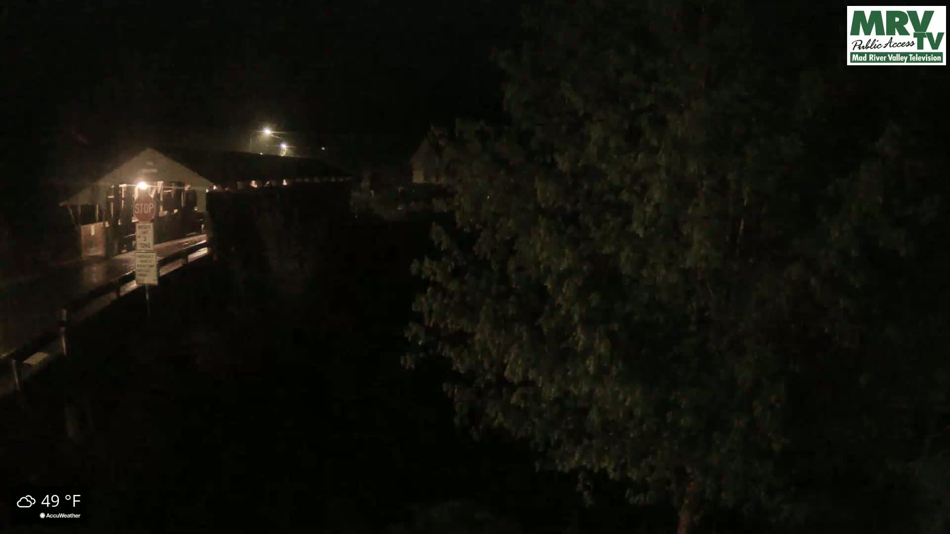 A nighttime view of a covered bridge during a rain shower, partially obscured by foliage.