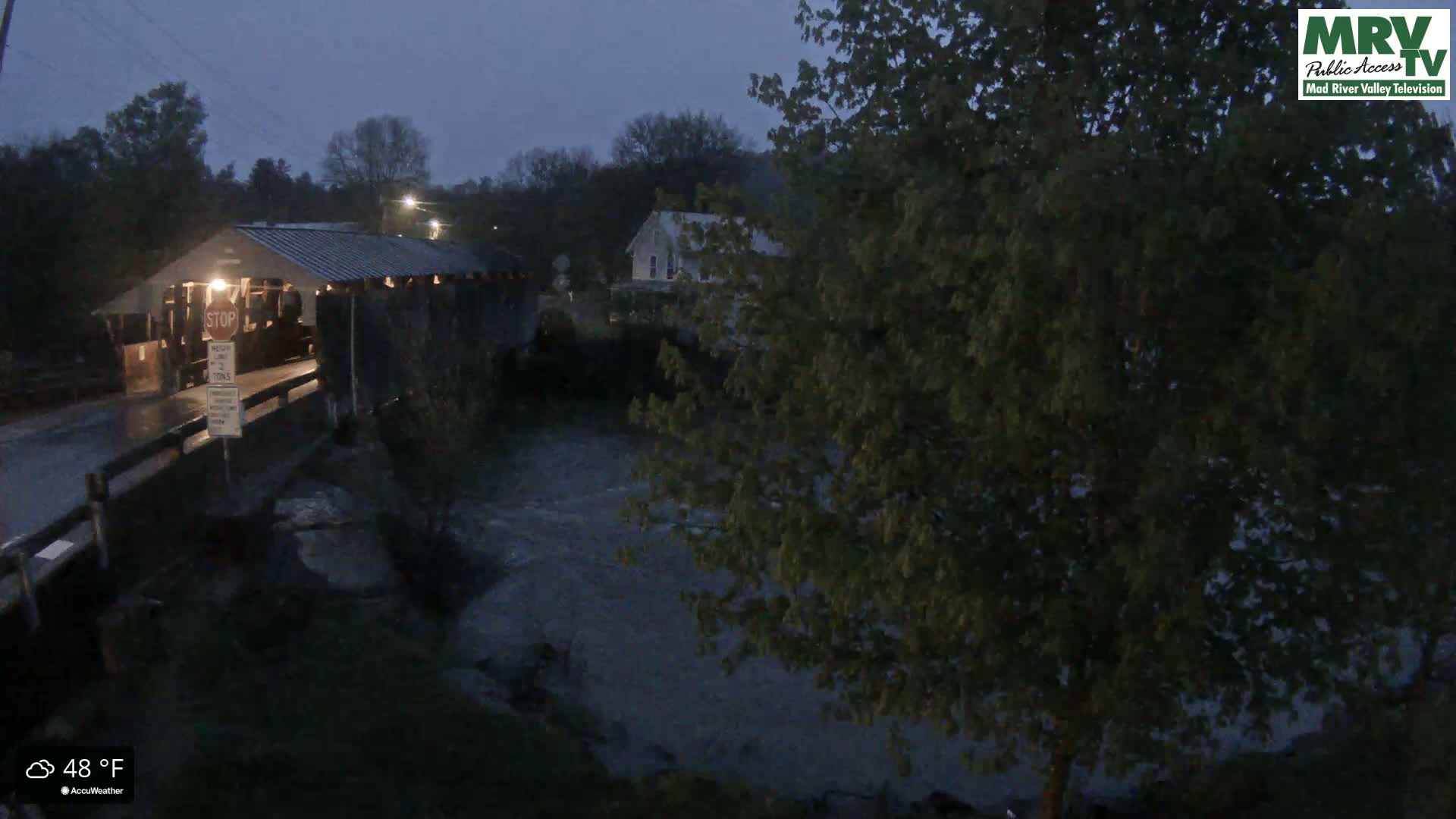 A covered bridge is visible in the rain at dusk, with a river flowing underneath and trees surrounding the area.