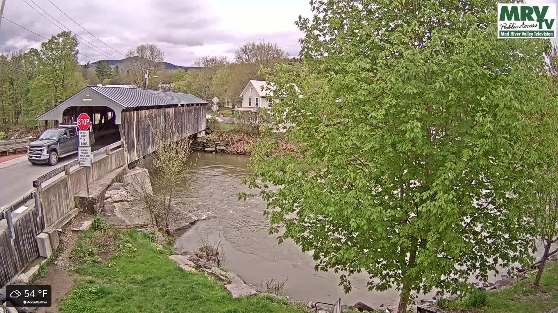 A covered bridge over a river is seen under cloudy skies, with a pickup truck driving across it and green trees and a house visible in the background.