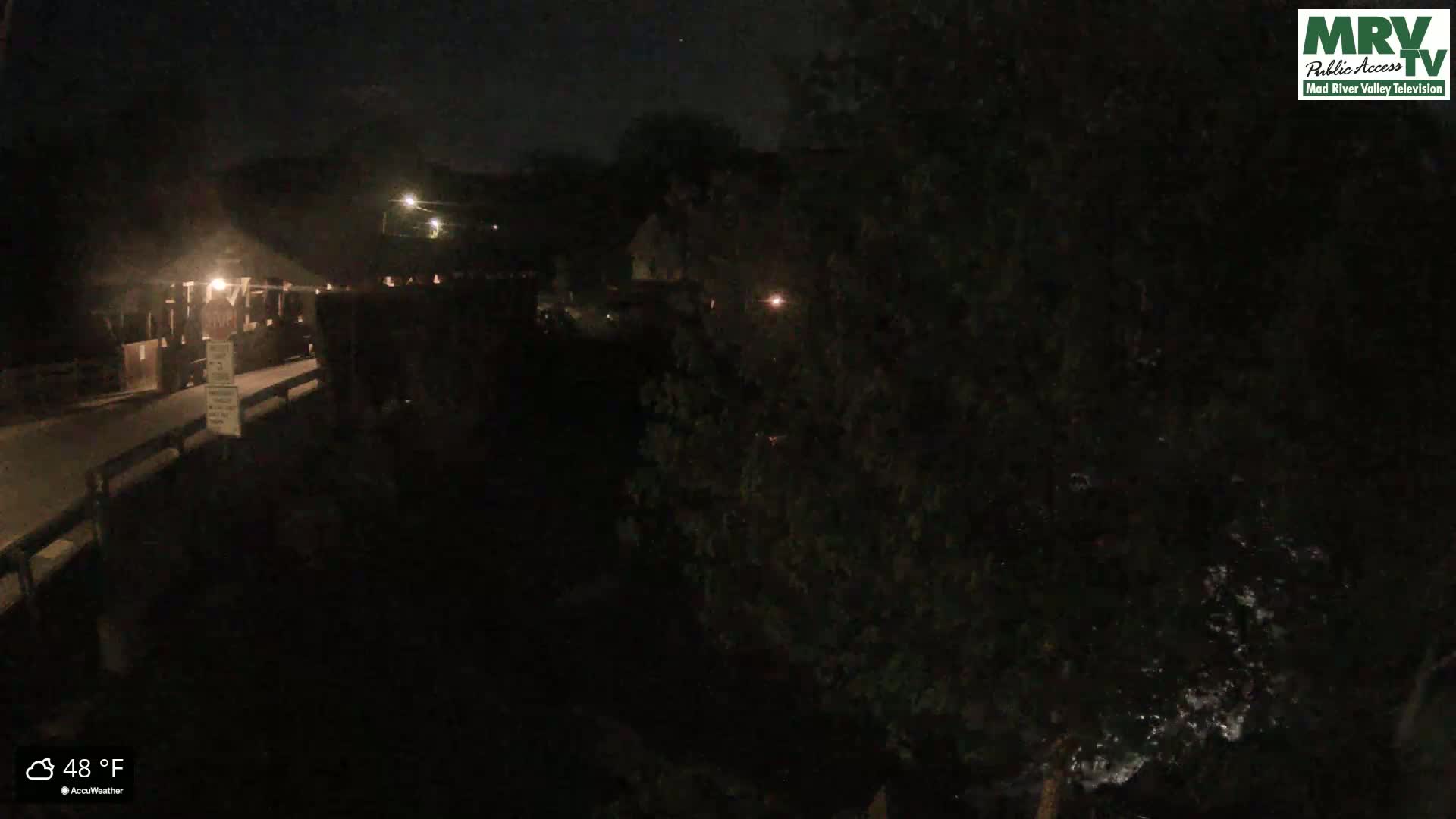 A nighttime view shows a bridge and a dark, tree-lined riverbank under a cloudy, 48-degree Fahrenheit sky.