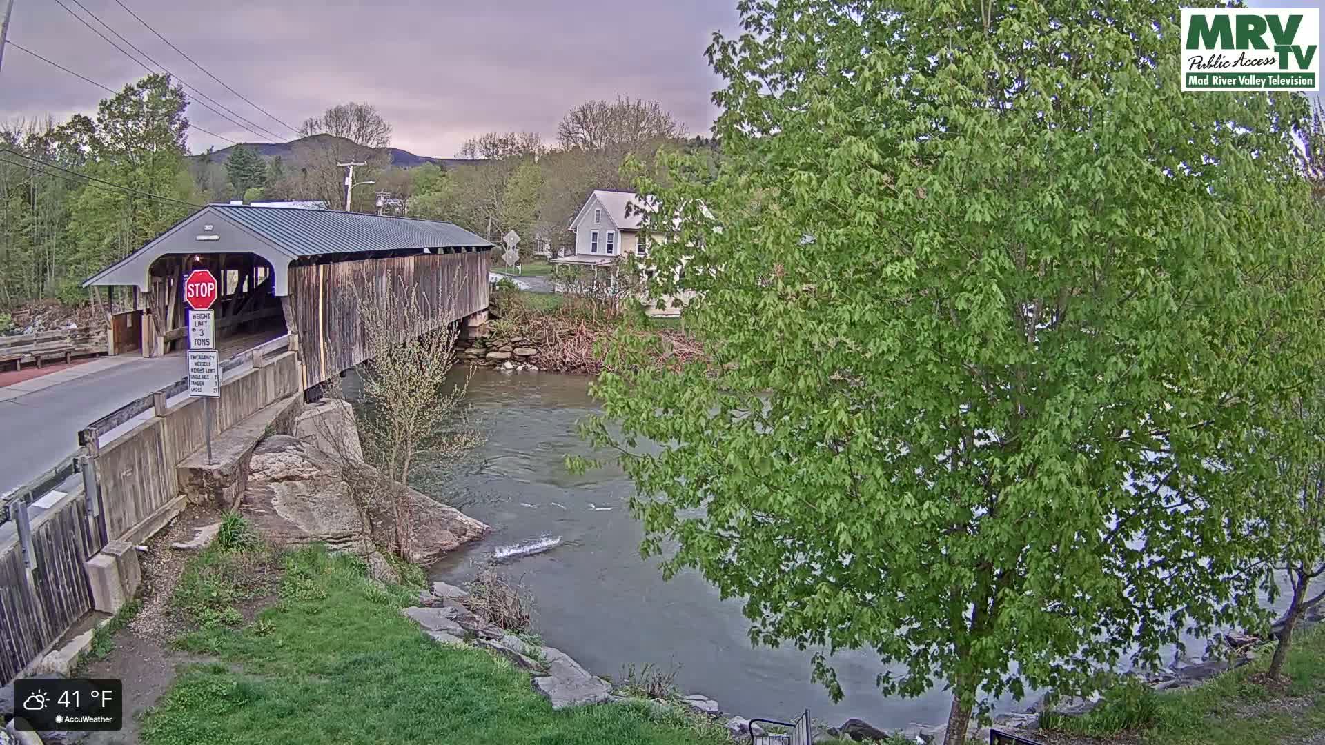 A covered bridge spans a flowing river under an overcast sky, with lush green trees and a house visible in the background.