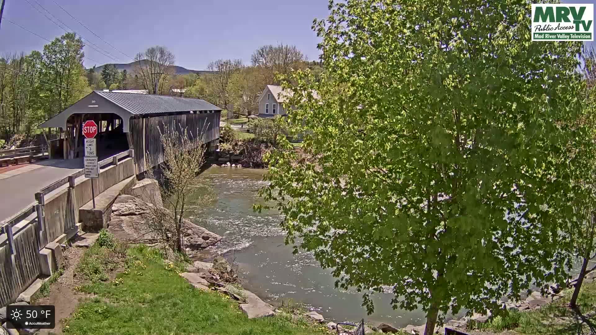 A covered bridge spans a flowing river under a sunny sky, with lush green trees and a house visible in the background.