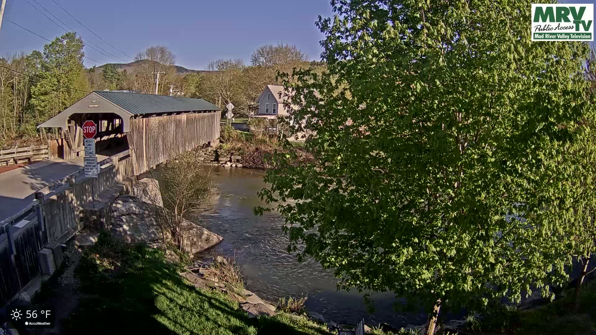 A wooden covered bridge spans a river on a sunny day, with lush green trees and a building visible in the background.