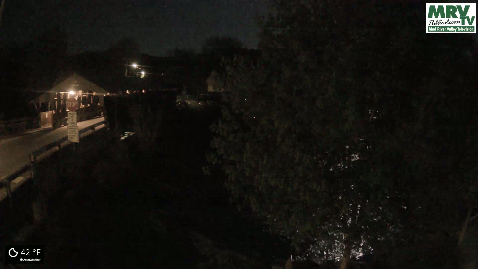 A nighttime view of a covered bridge over a dark body of water, under a clear sky at 42°F.
