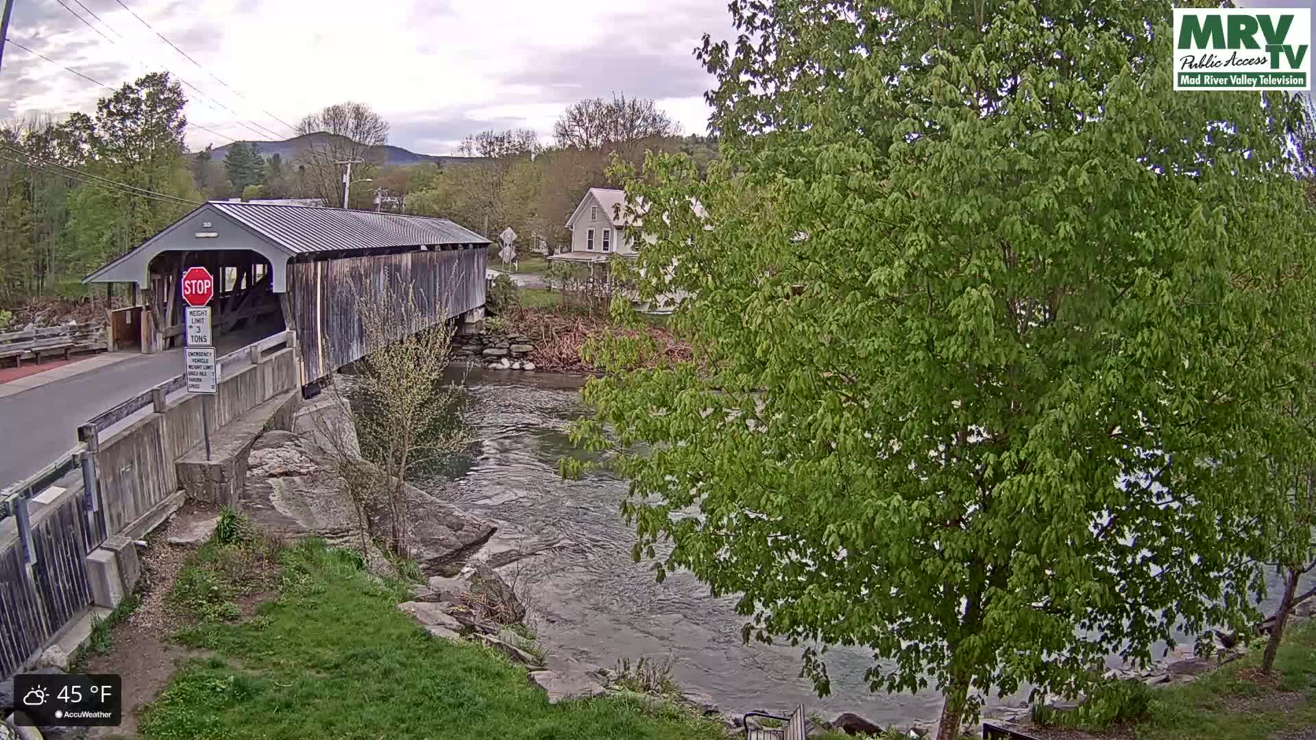 A covered bridge spans a river under an overcast sky, with lush green trees and a house visible in the background.