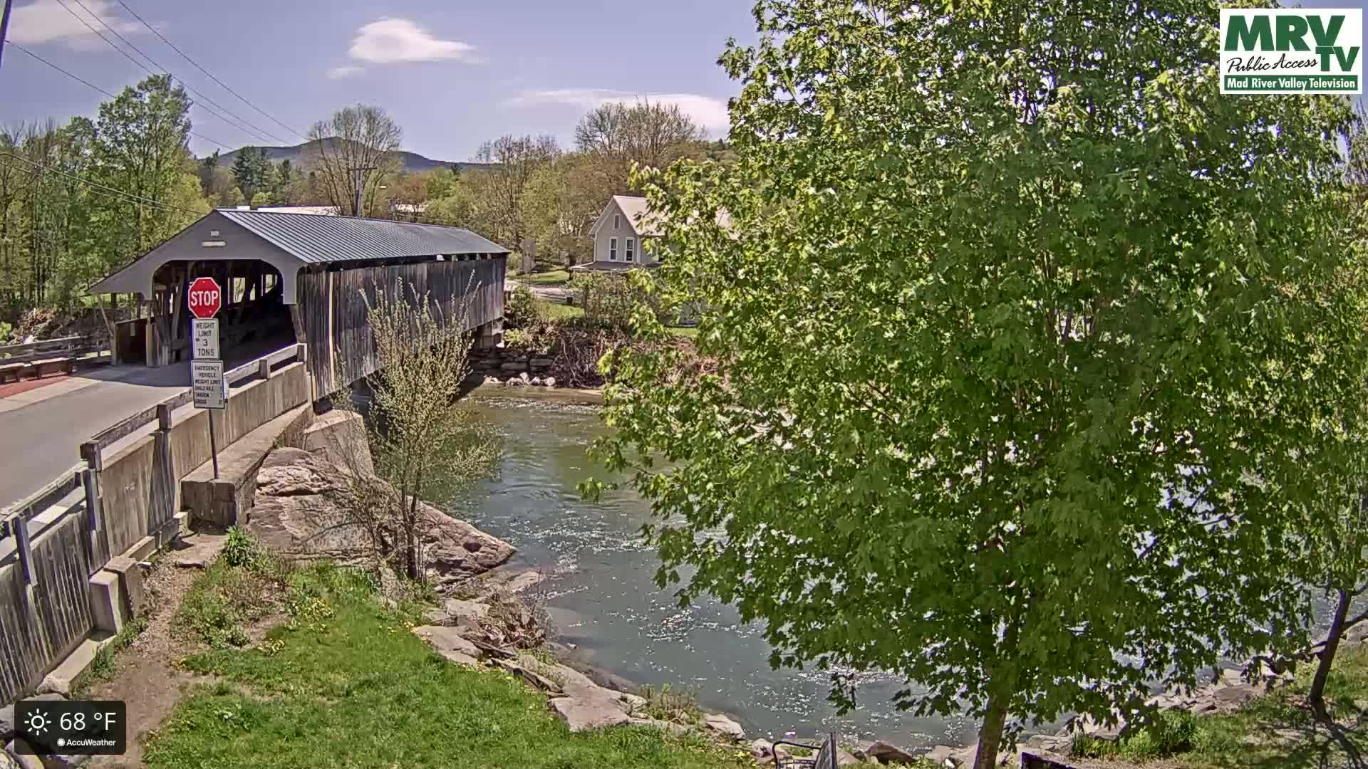 A covered bridge spans a river under a partly sunny sky, with lush green trees and a house visible in the background.