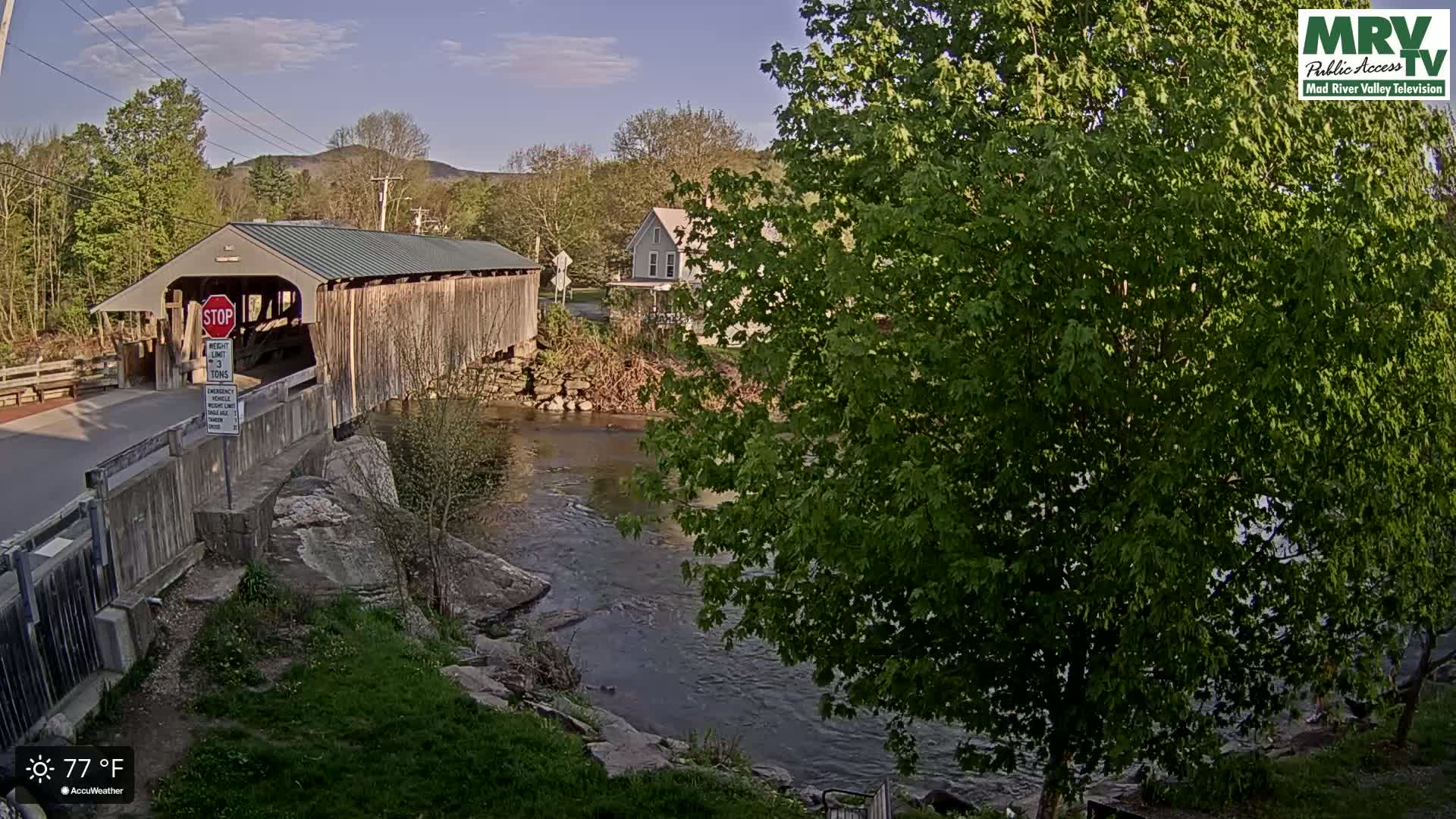 A covered wooden bridge spans a calm river under a clear, sunny sky, with lush green trees and a house visible in the background.