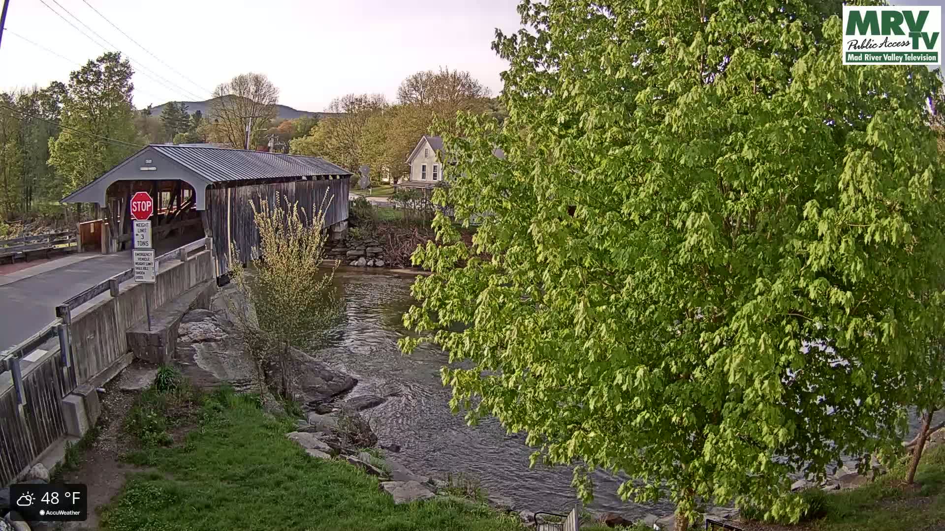 A covered bridge spans a river under a partly cloudy sky, with lush green trees and foliage surrounding the waterway.