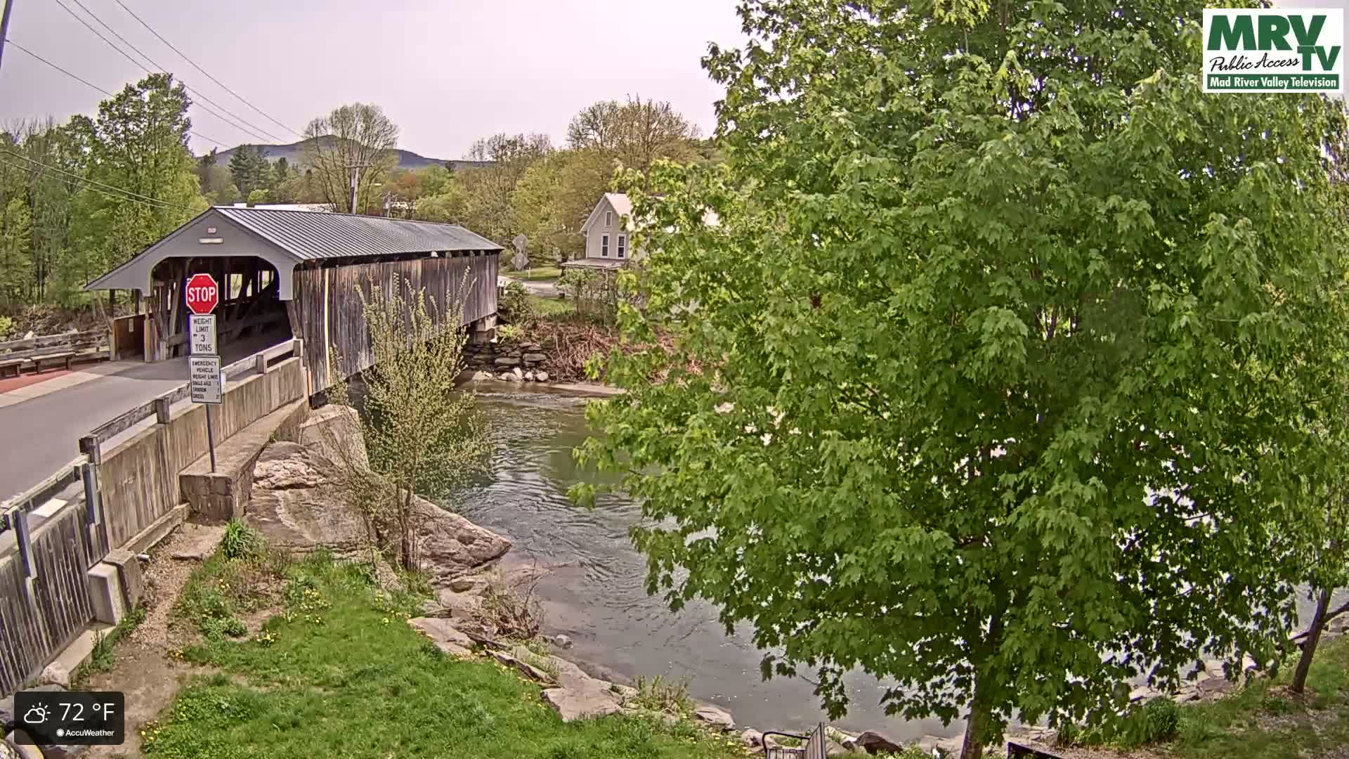 A wooden covered bridge spans a calm river under a partly cloudy sky on a 72°F day, with lush green trees and grassy banks surrounding the scene.
