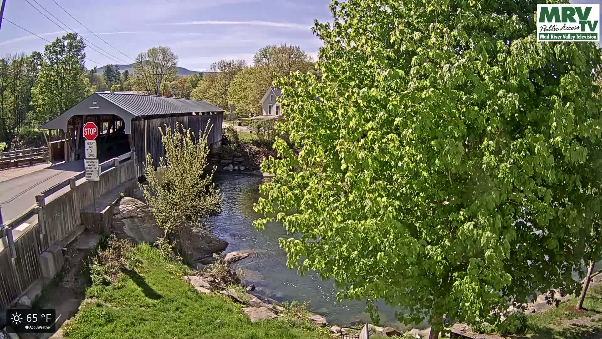 A covered bridge spans a river under a sunny sky, with lush green foliage surrounding the waterway.