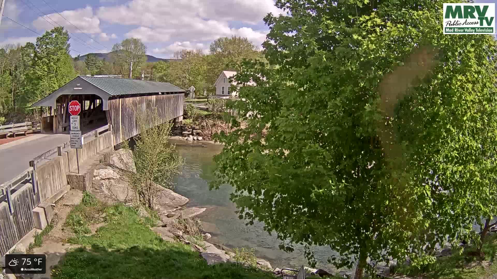 A covered bridge spans a river under a partly cloudy sky, with lush green trees and foliage surrounding the scene.