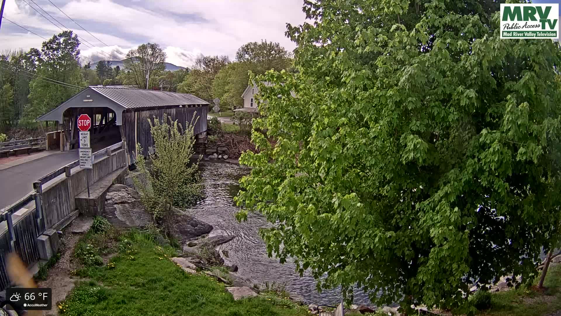 A covered bridge spans a river under a partly cloudy sky, with lush green trees and vegetation surrounding the area.