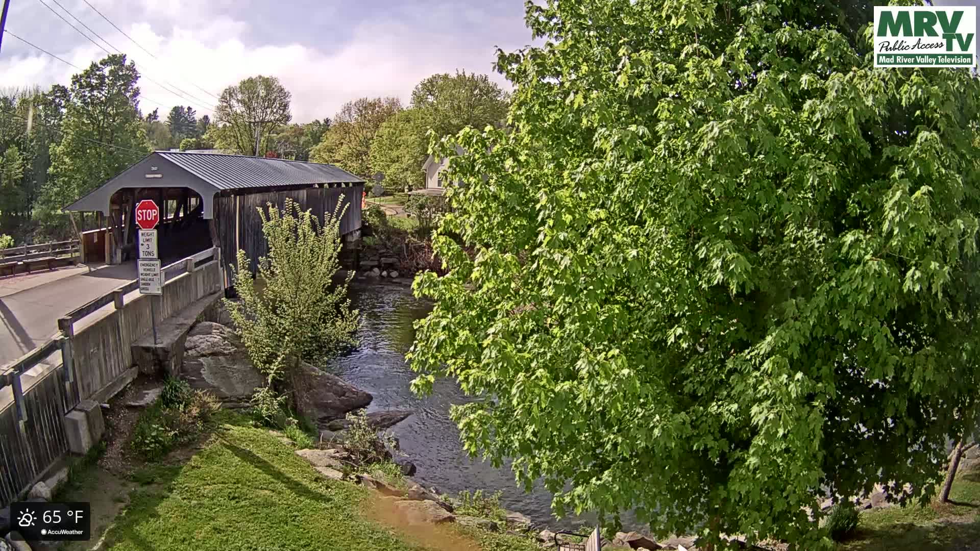 A covered bridge spans a river under a partly cloudy sky, with lush green foliage surrounding it.