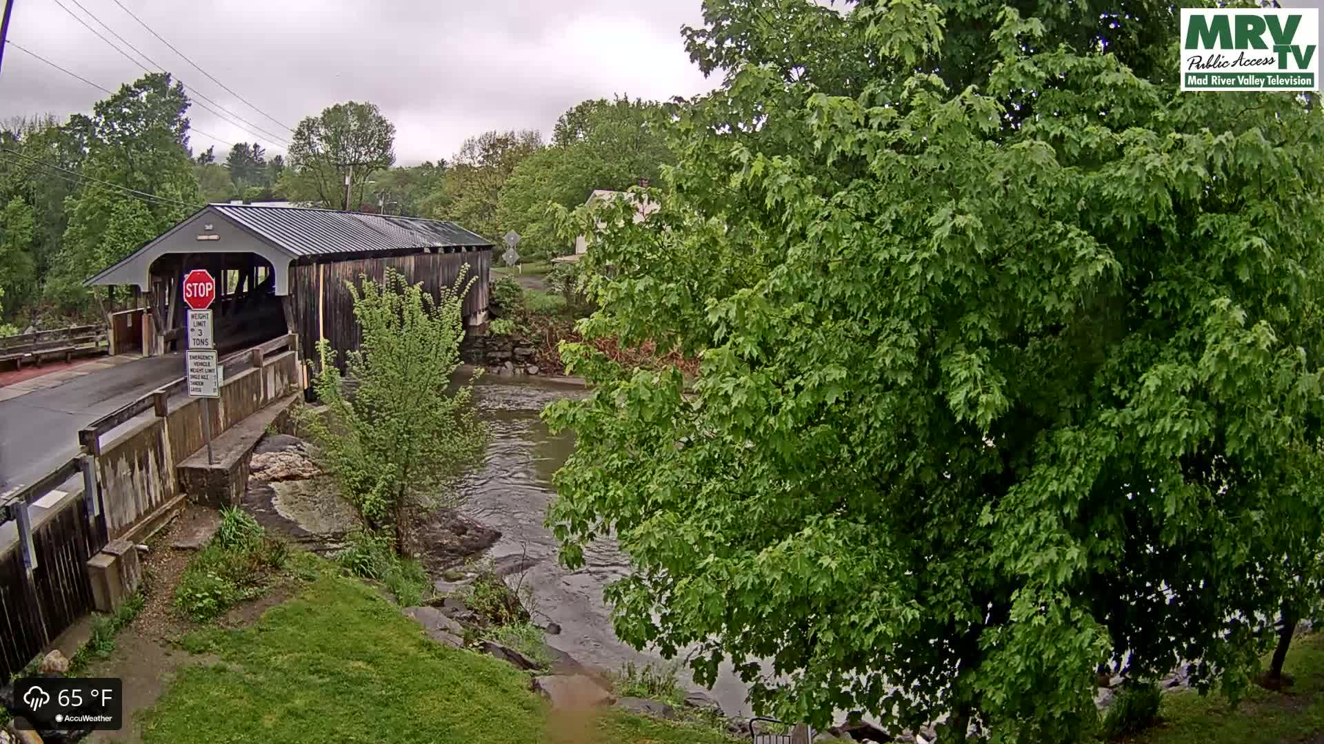 A covered bridge spans a river under an overcast sky, with lush green foliage surrounding the scene.