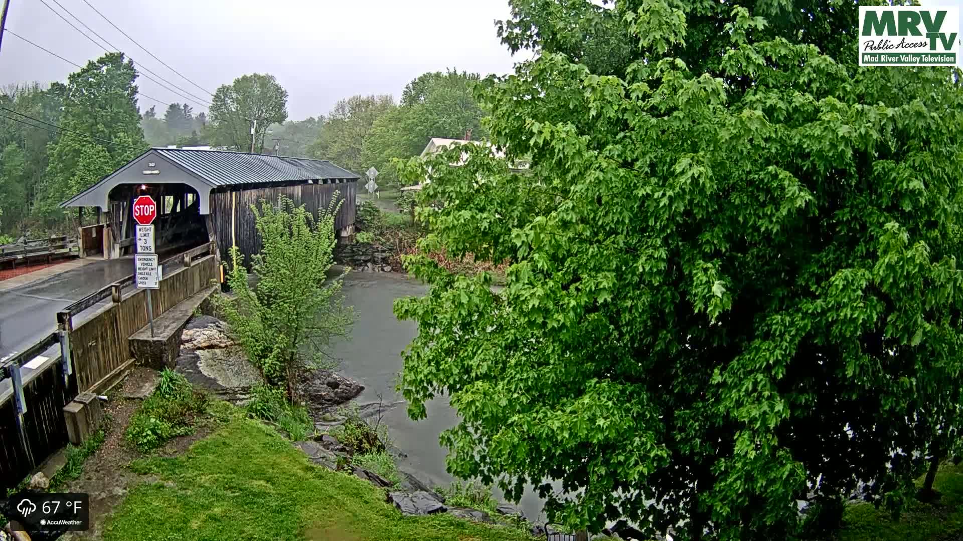 A covered bridge is situated beside a calm river under a rainy sky, with lush green trees surrounding the scene.