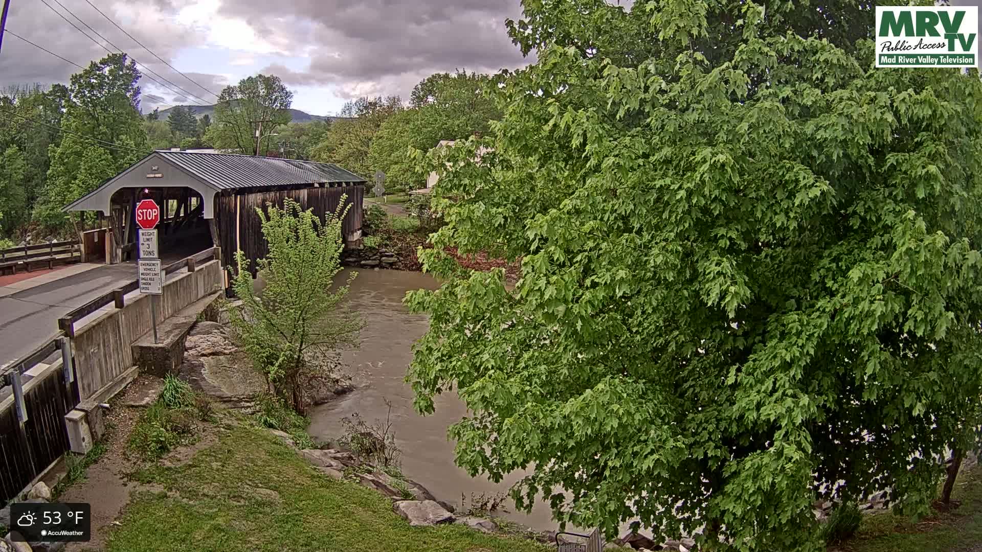 A covered bridge spans a muddy river under a cloudy sky, with lush green foliage surrounding the scene.