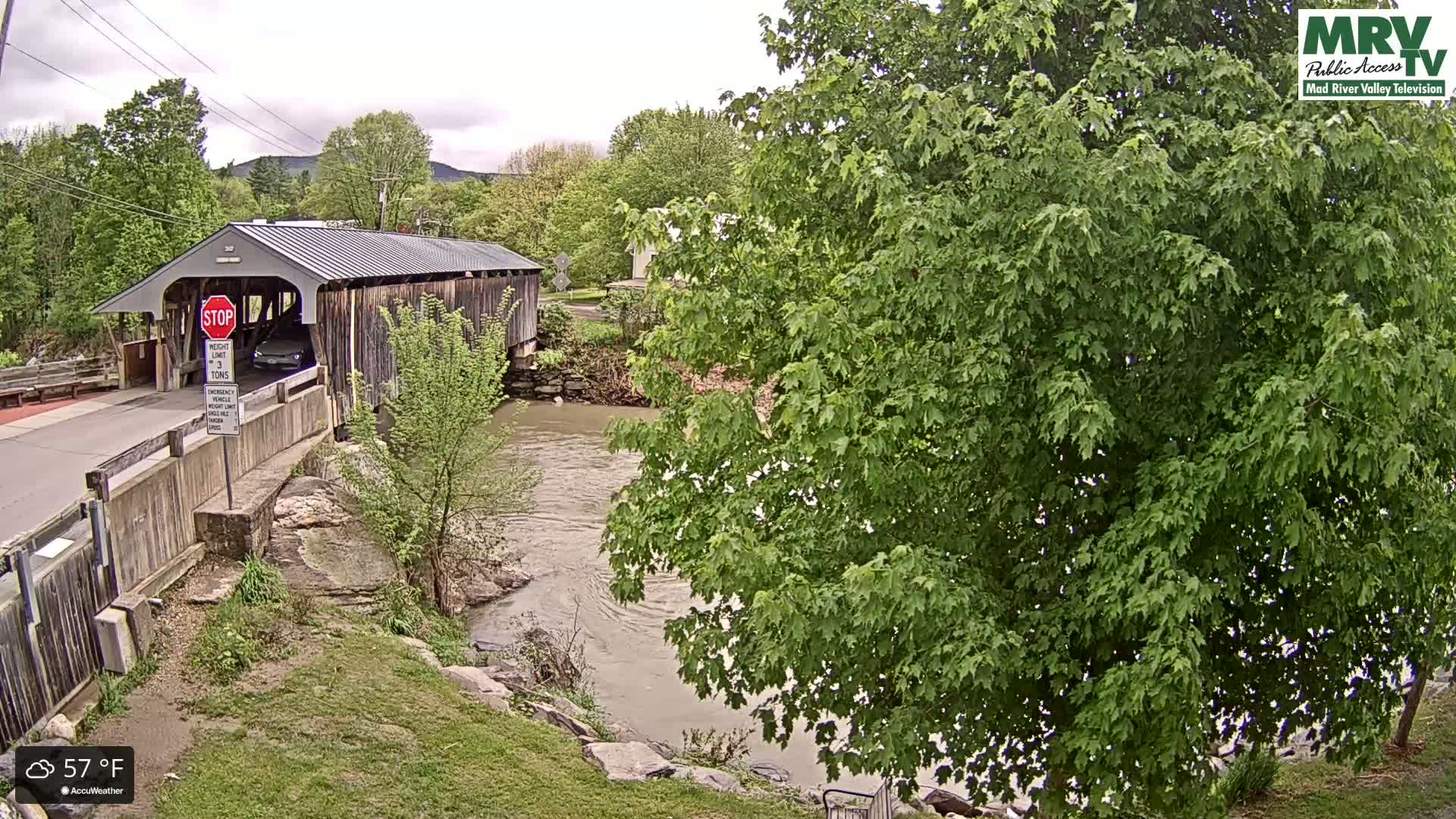 A covered bridge spans a muddy river under a partly cloudy sky; lush green trees and grass surround the scene.