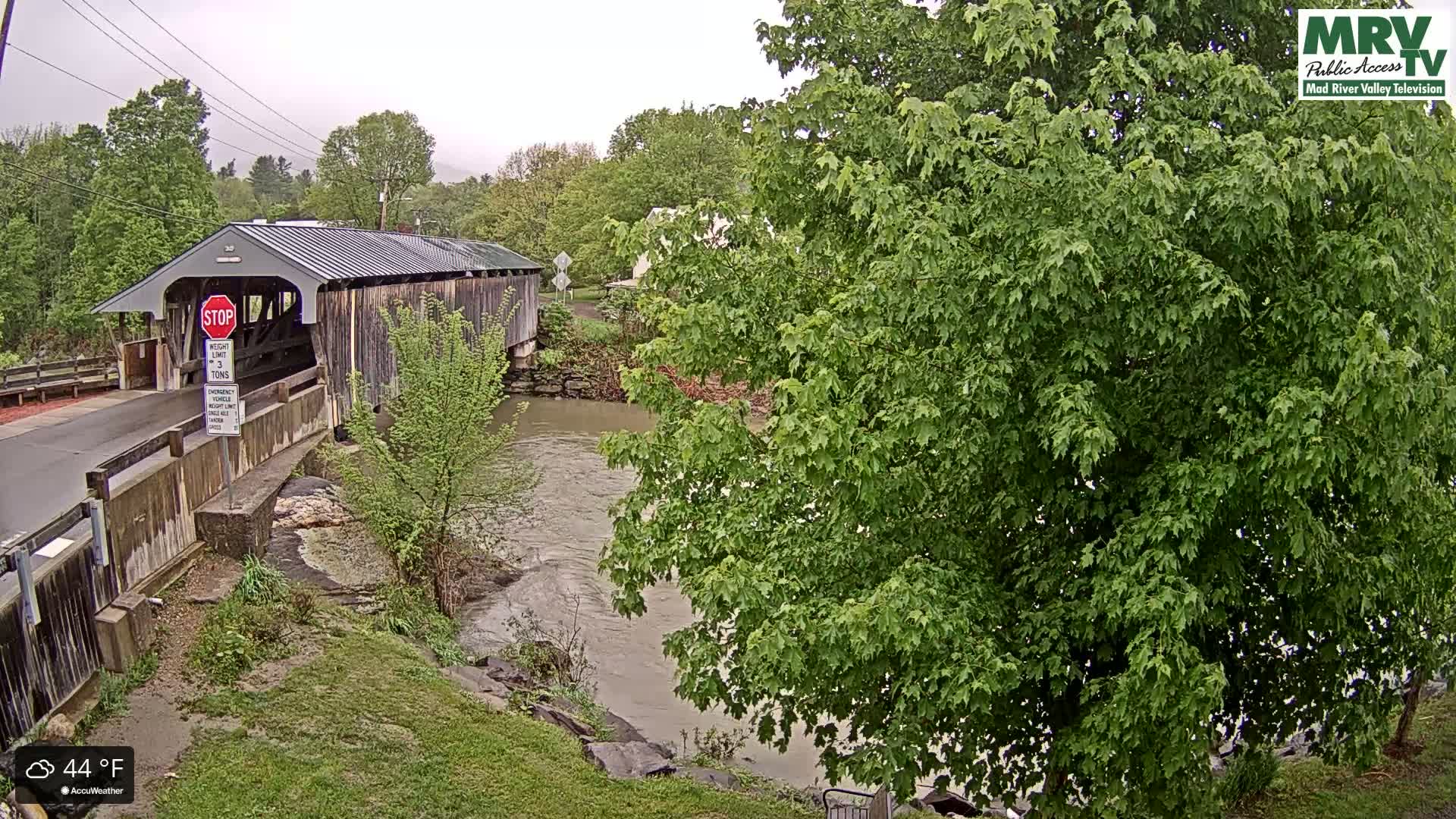 A covered wooden bridge spans a muddy river under an overcast sky.