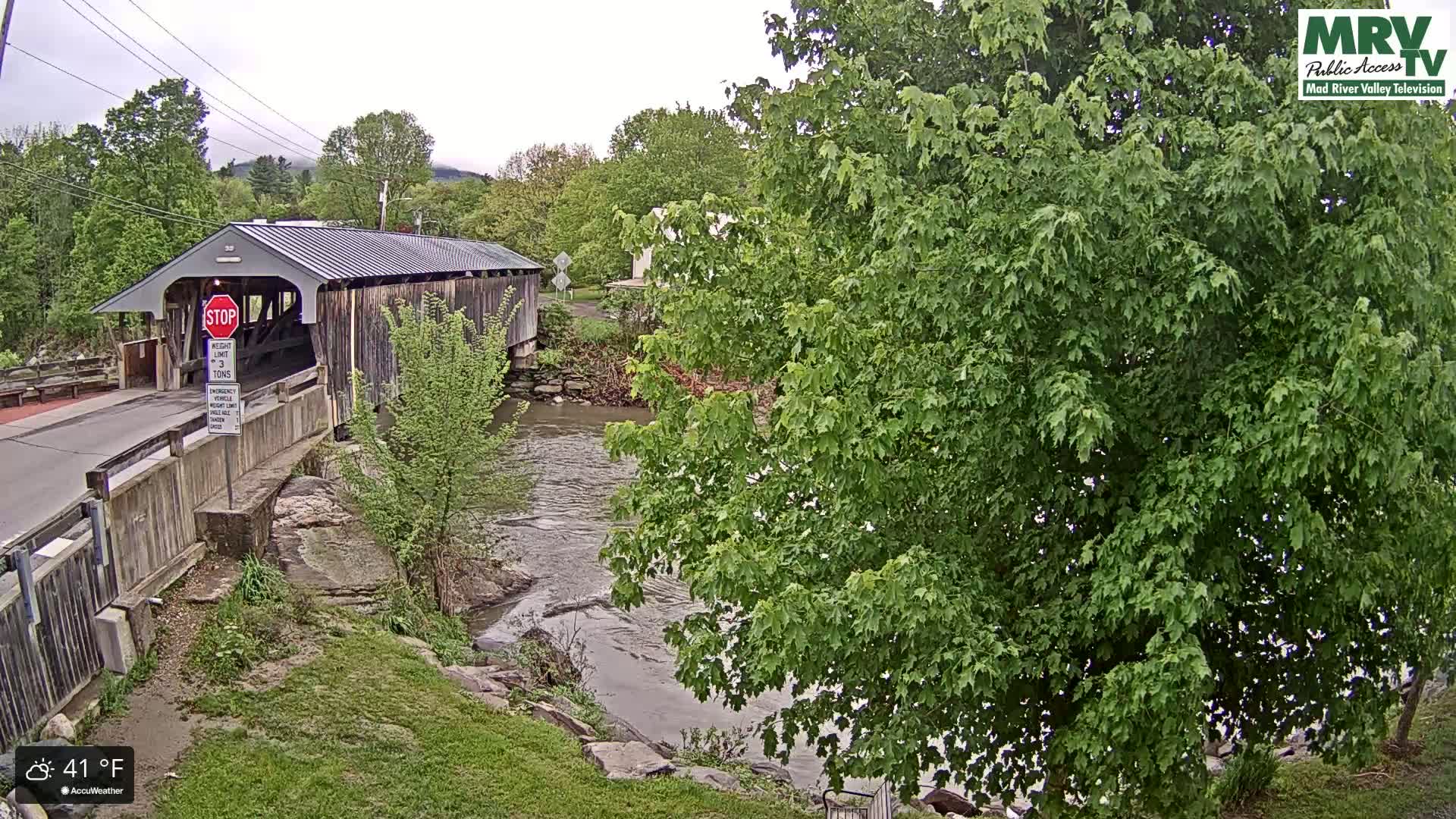 A covered bridge spans a river under an overcast sky, with lush green trees lining the banks.