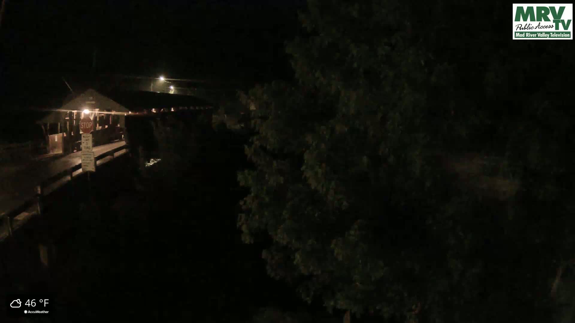 A dark, nighttime view of a covered bridge, partially obscured by trees, under cloudy skies with a temperature of 46°F.