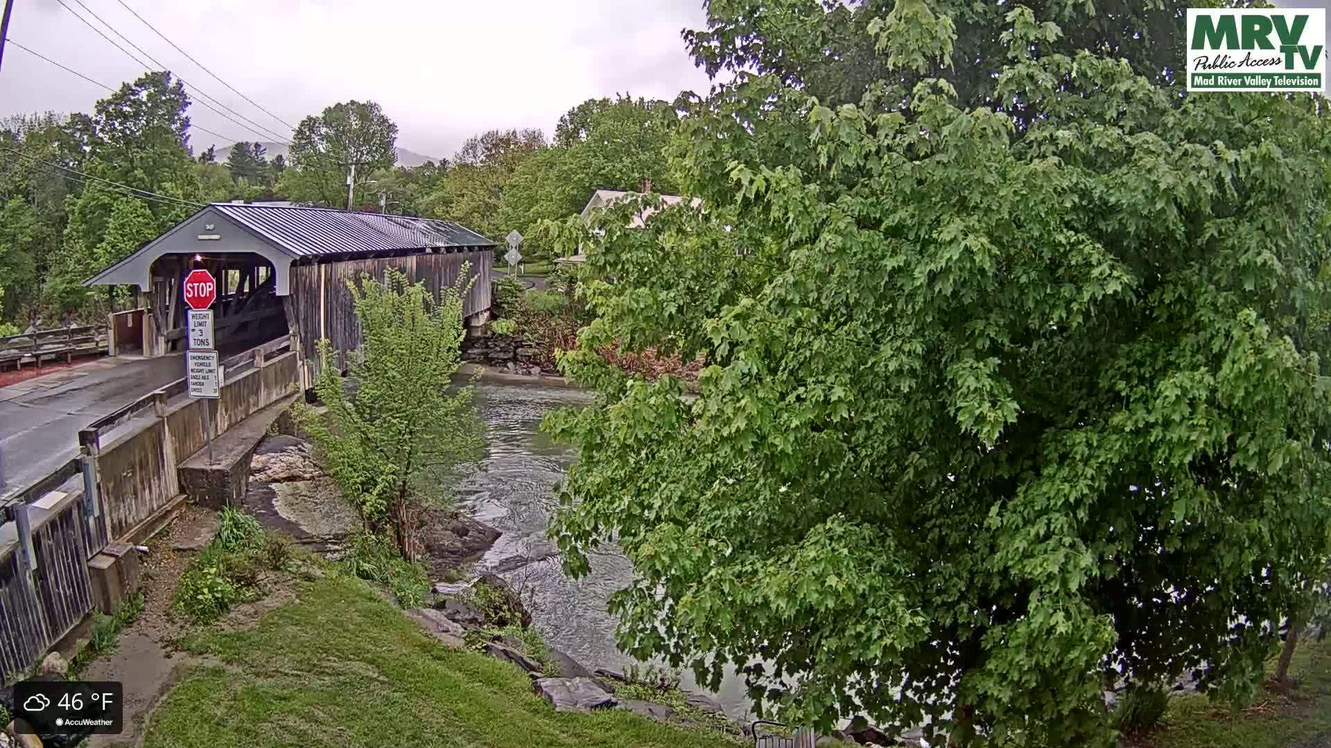 A covered bridge spans a flowing river under an overcast sky.
