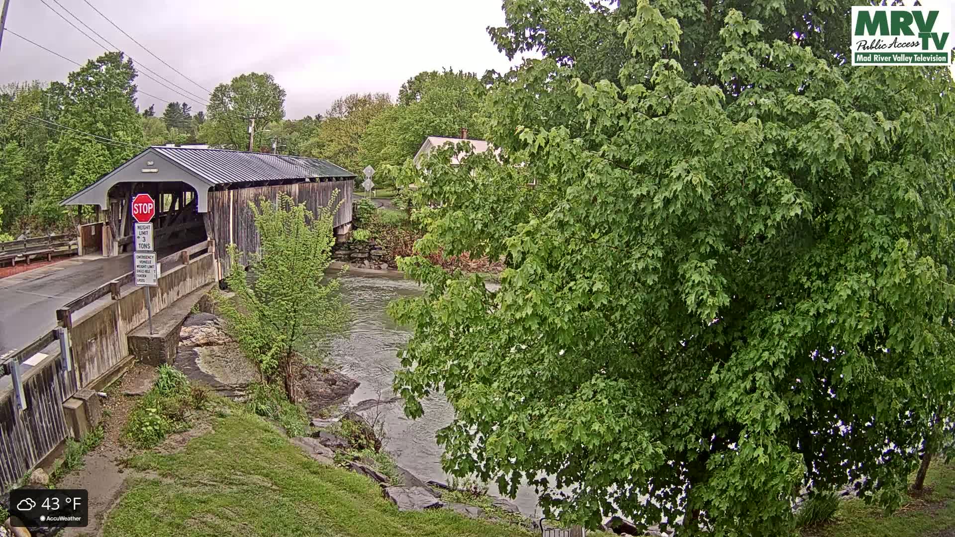A covered bridge spans a river under an overcast sky, with lush green foliage surrounding the scene.