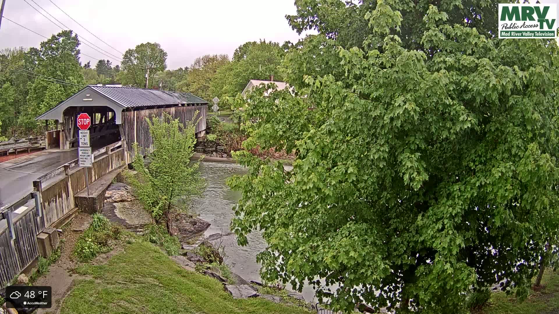 A covered bridge spans a calm river under an overcast sky, surrounded by lush green trees and grassy banks.