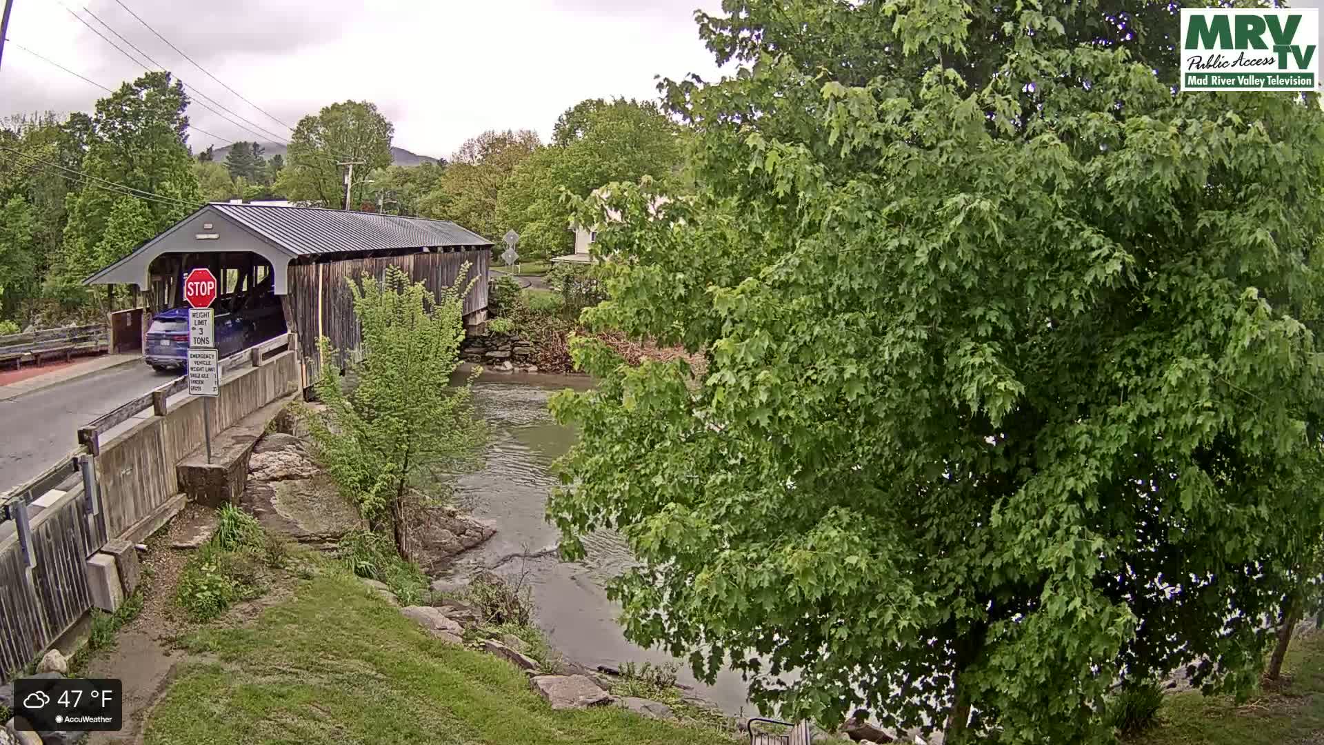 A covered bridge spans a calm river under an overcast sky, with lush green foliage surrounding the scene.