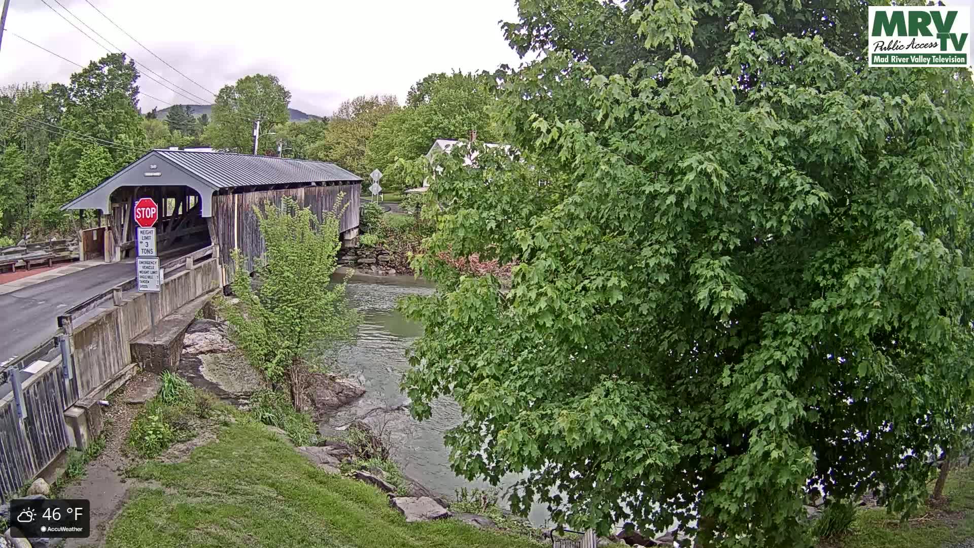 A covered bridge spans a calm river under an overcast sky, surrounded by lush green foliage.