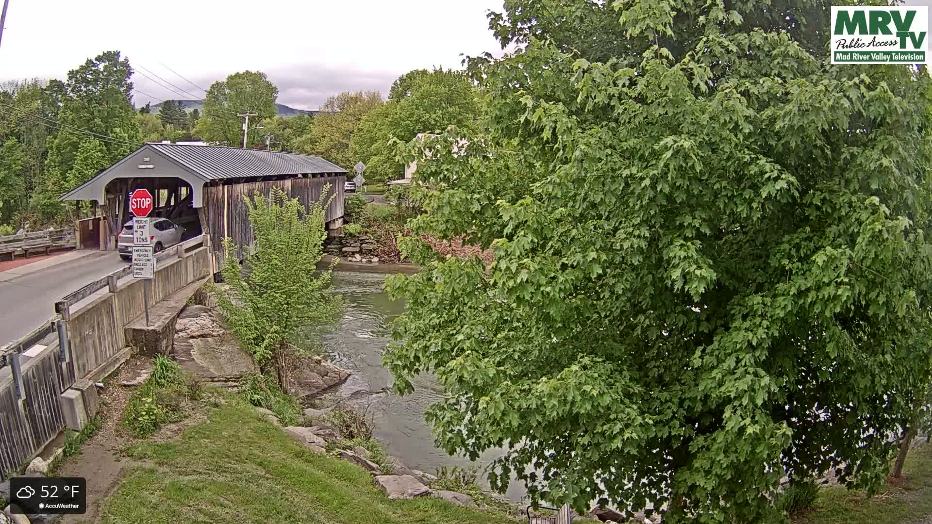 A covered bridge spans a river under overcast skies, with a car driving across it.