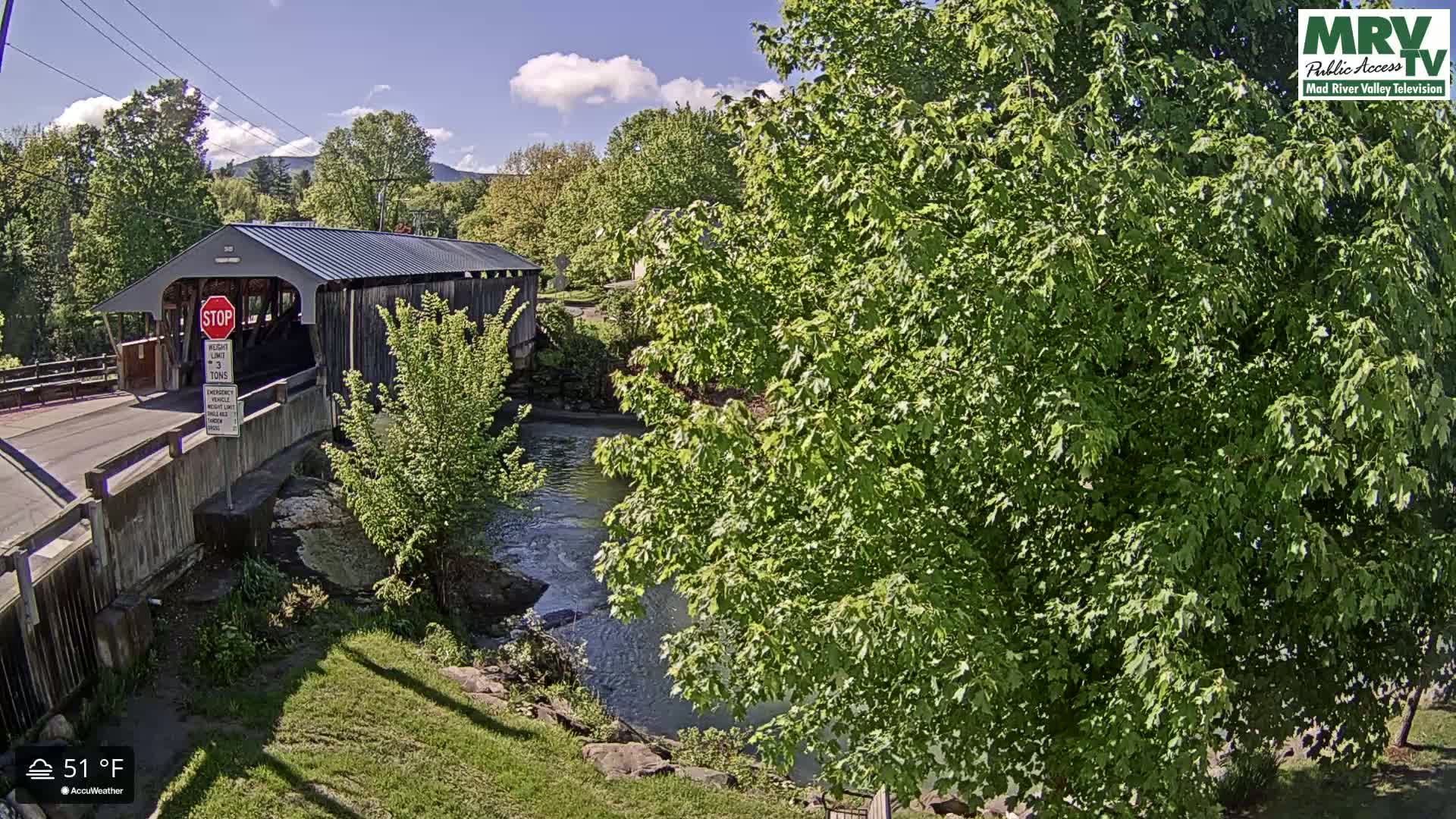 A covered bridge spans a river under a partly cloudy sky, with lush green foliage surrounding the waterway.