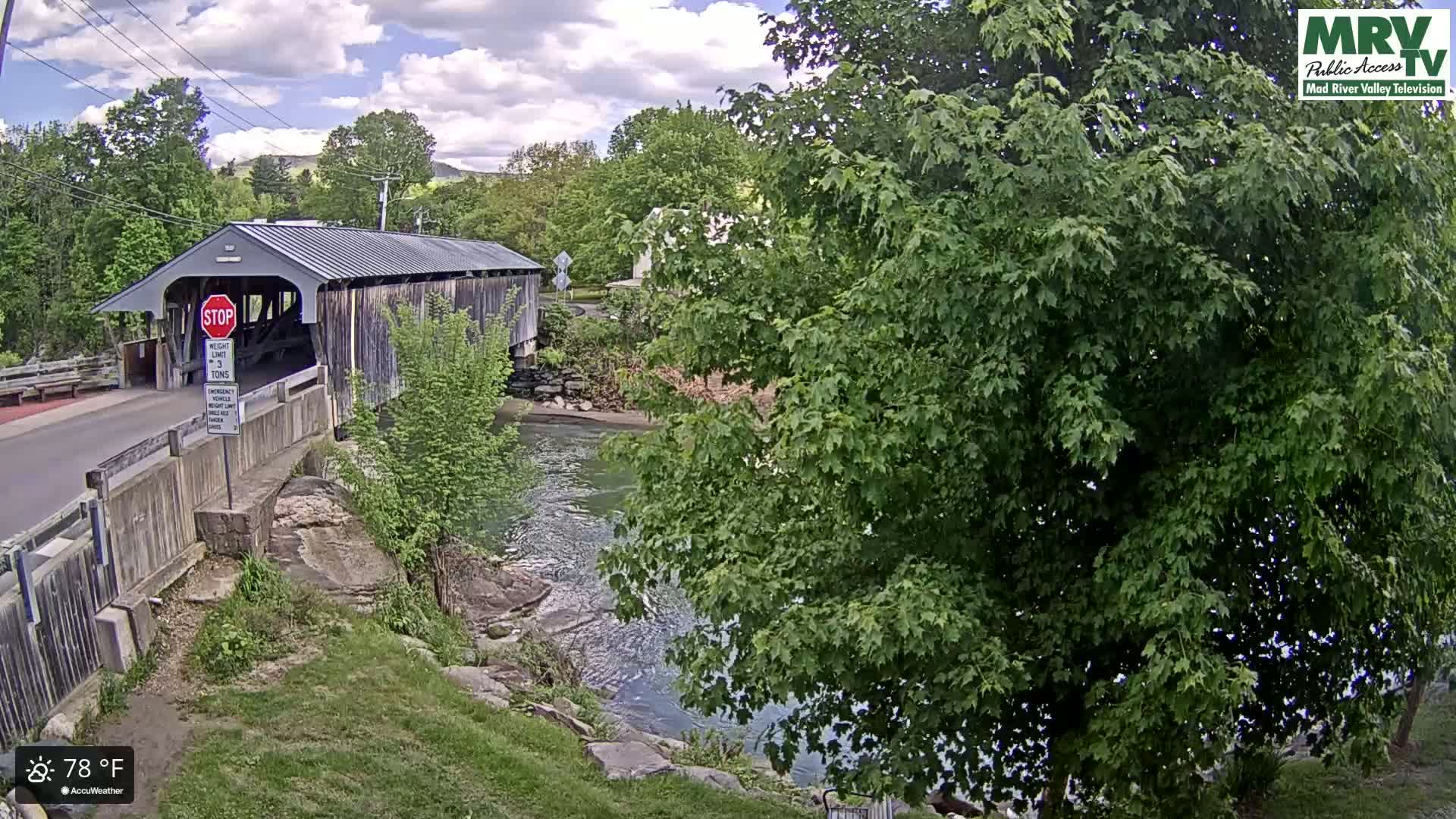 A covered bridge spans a river under a partly cloudy sky, with lush green trees and grassy banks surrounding the water.