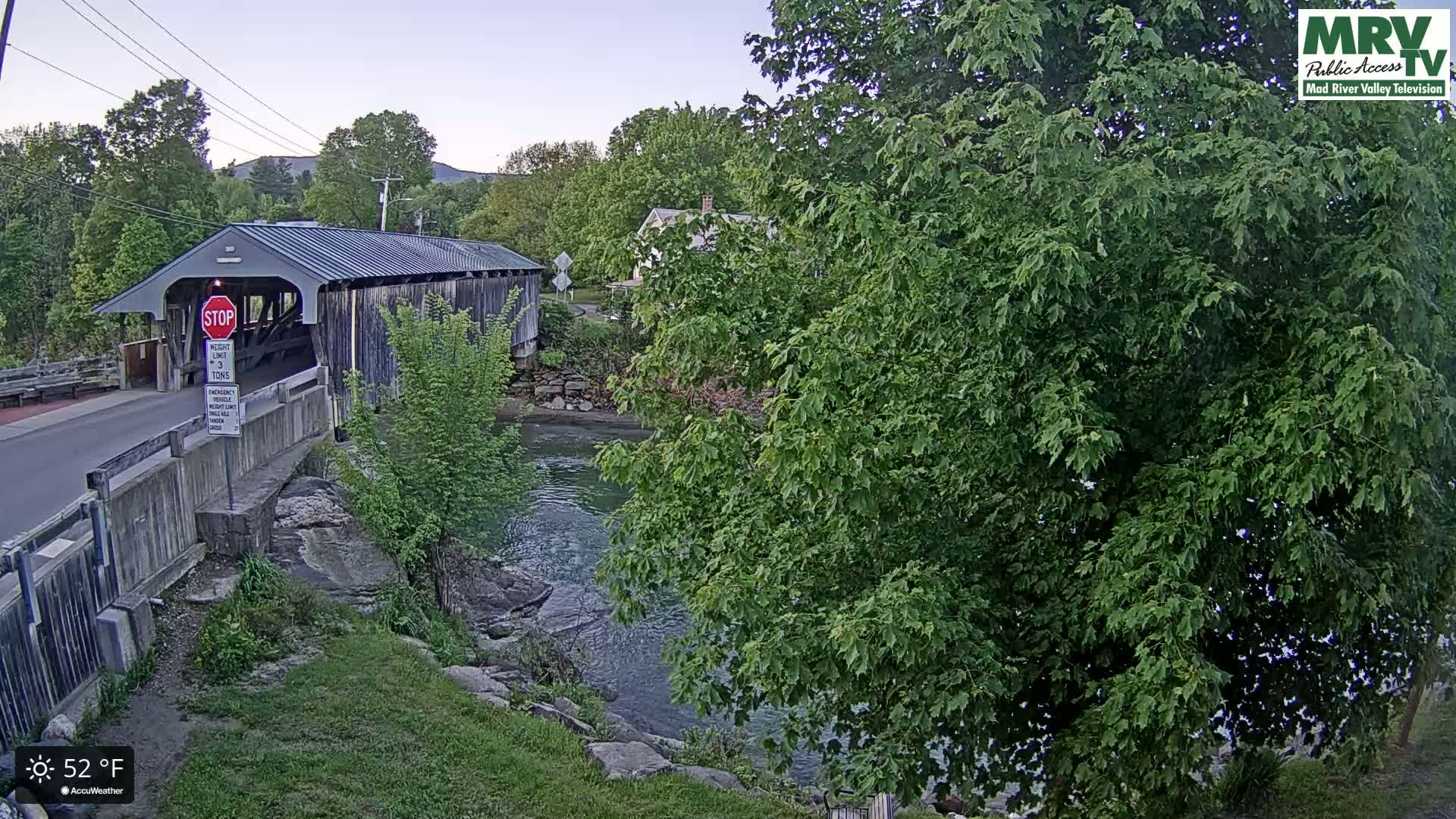 A covered bridge spans a calm river under a clear, twilight sky, surrounded by lush green trees and foliage.