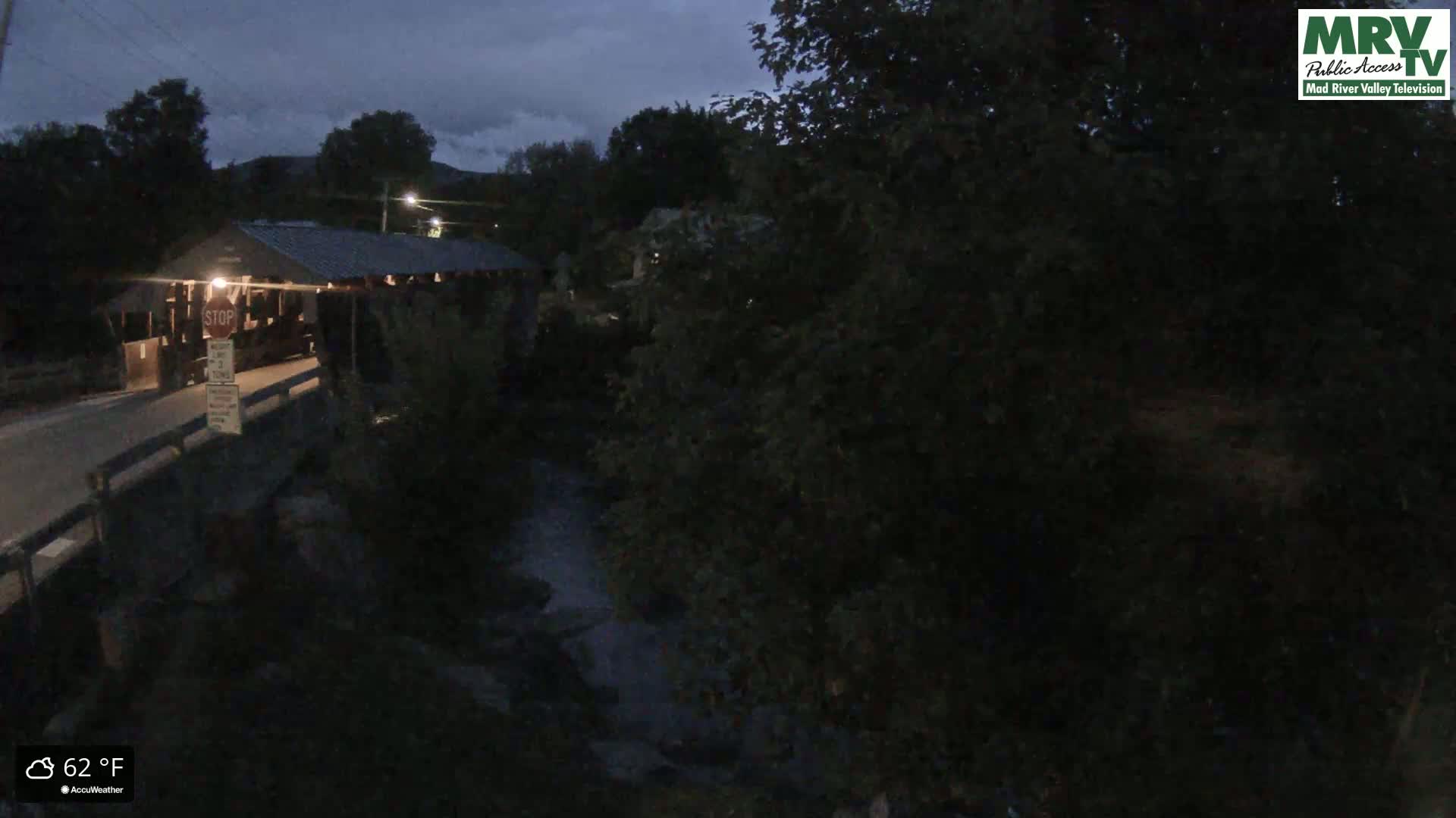A covered bridge sits over a dark stream under a cloudy night sky.
