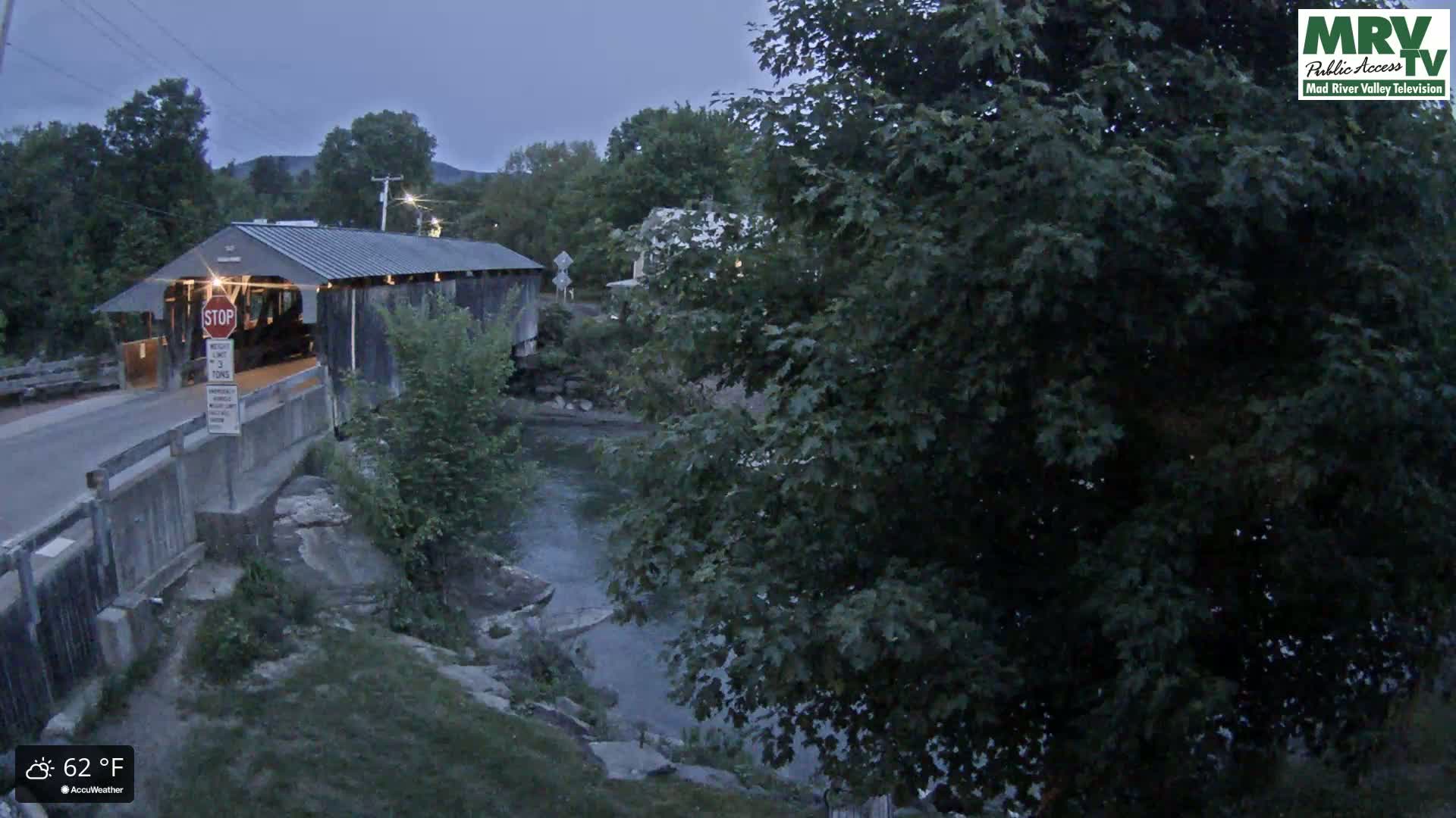 A covered bridge stands over a calm river at dusk, surrounded by lush green trees under a cloudy sky.