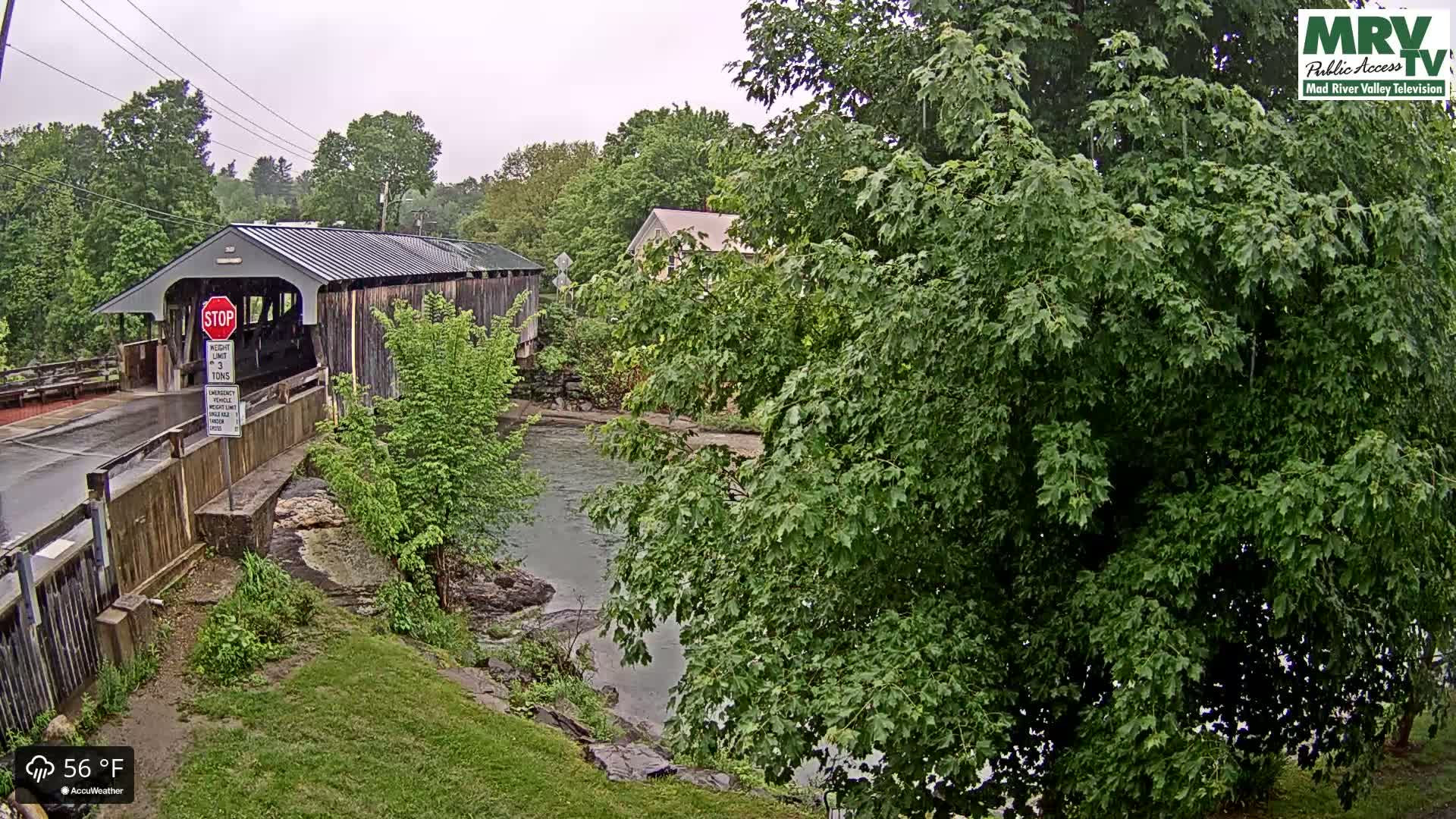 A covered bridge spans a calm river under an overcast sky, with lush green foliage surrounding the scene.