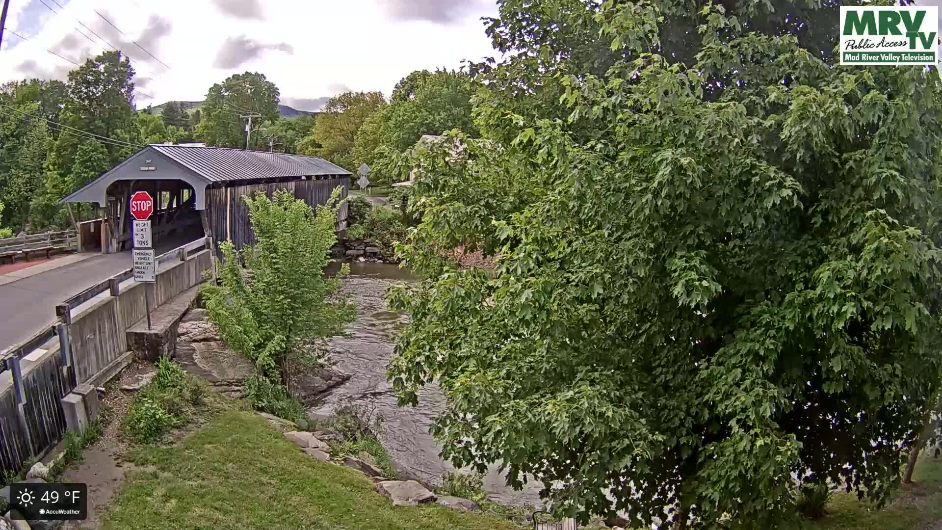 A covered bridge spans a flowing river under a partly cloudy sky, surrounded by lush green trees and vegetation.