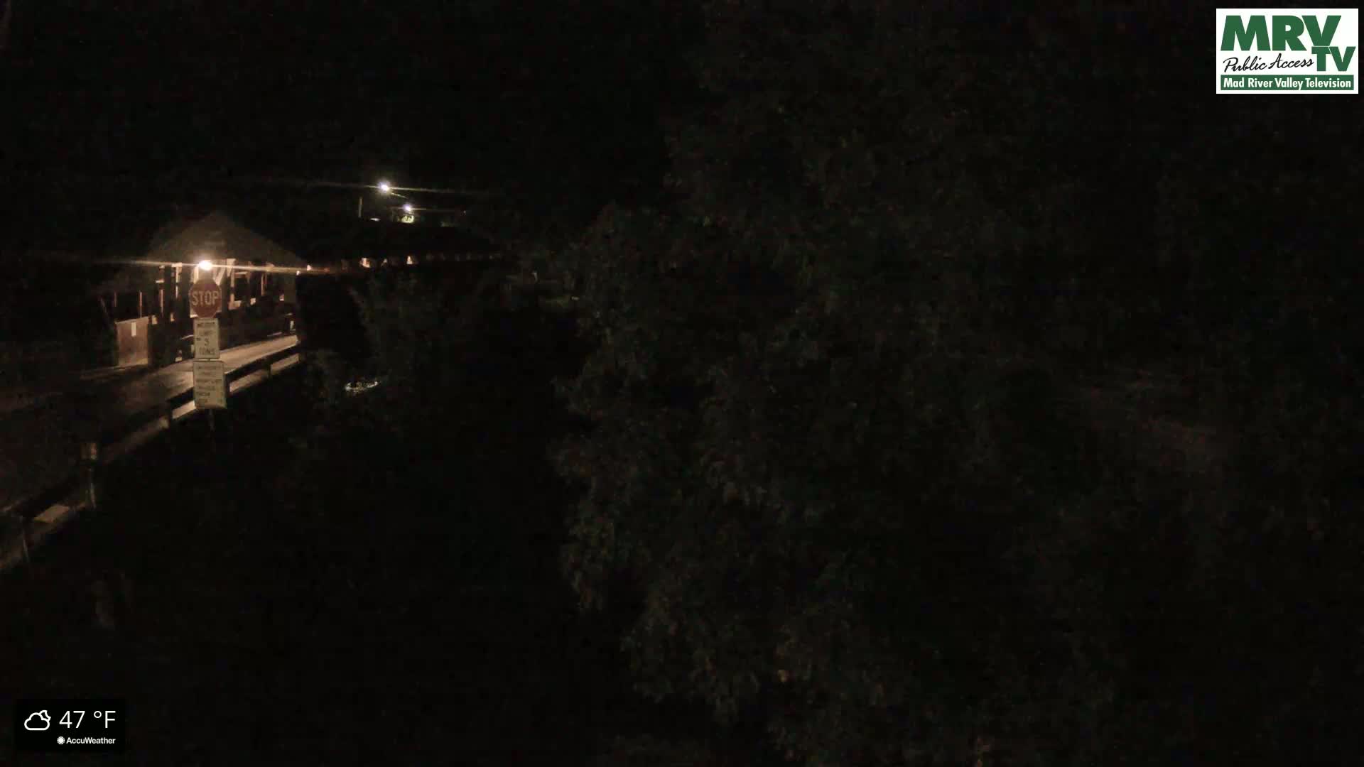 A nighttime, low-angle view shows a dark, empty road leading to a covered bridge, with dark foliage obscuring much of the background, and the temperature is 47°F.