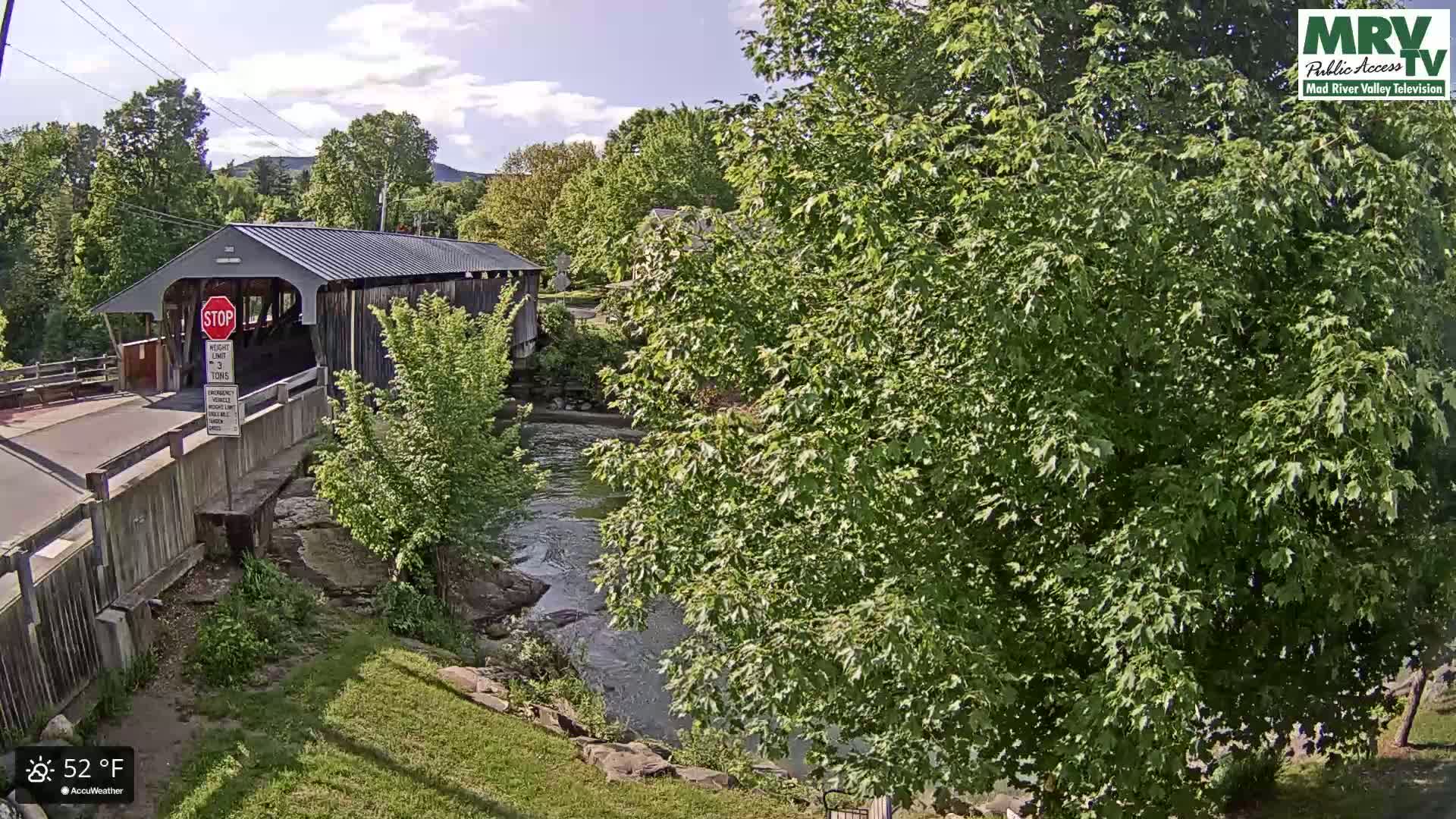 A covered bridge spans a river under a partly cloudy sky, with lush green trees lining the banks.