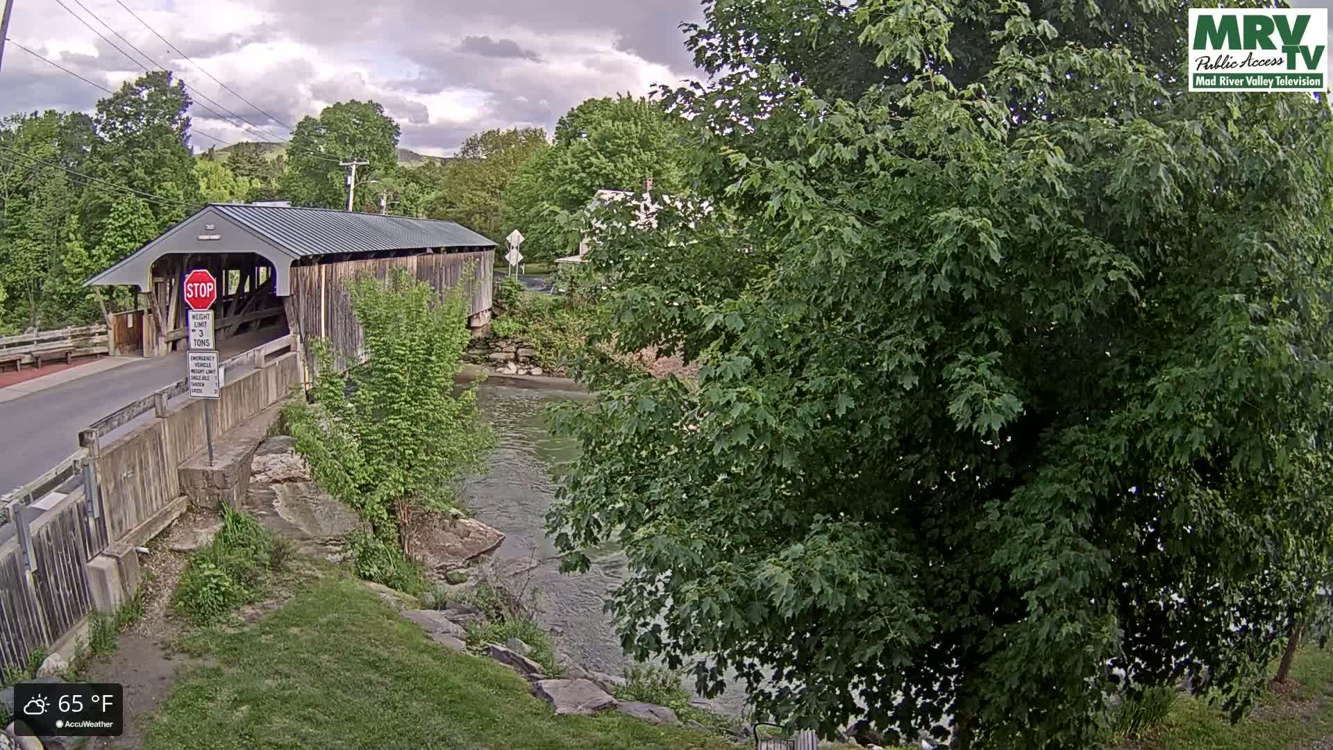 A covered bridge spans a river under a partly cloudy sky, with lush green trees lining the riverbanks and surrounding the area.