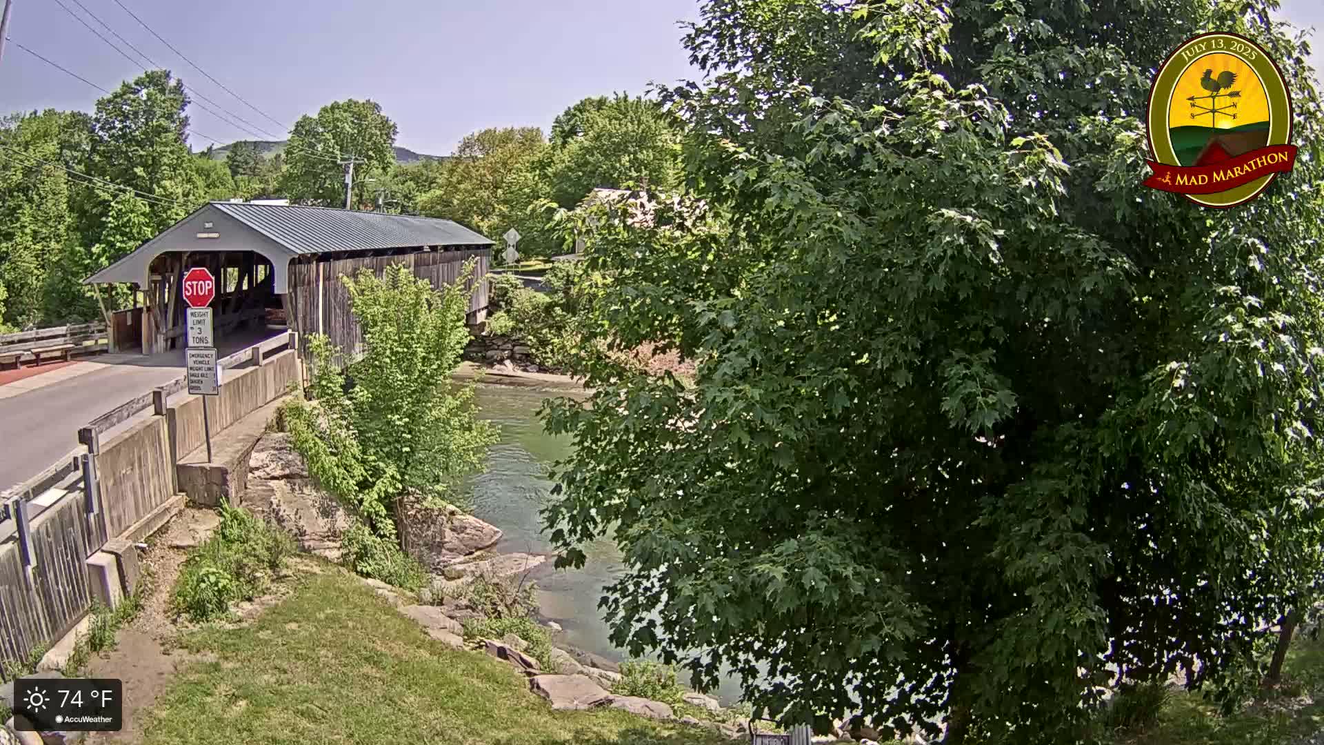 A covered bridge spans a calm river on a sunny 74°F day, with lush green trees and foliage surrounding the scene.