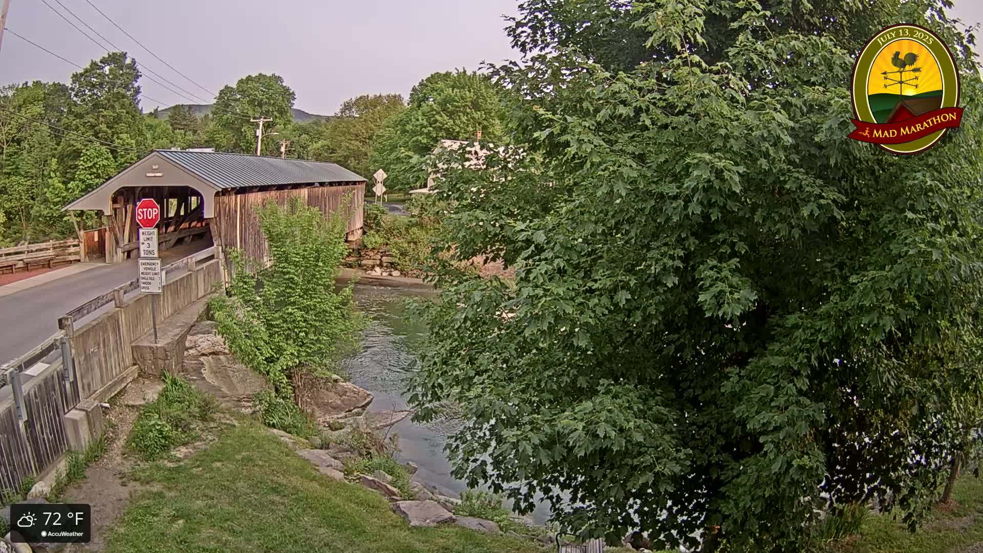 A covered bridge spans a river bordered by lush green trees and rocky banks under a partly cloudy sky; the temperature is 72°F.