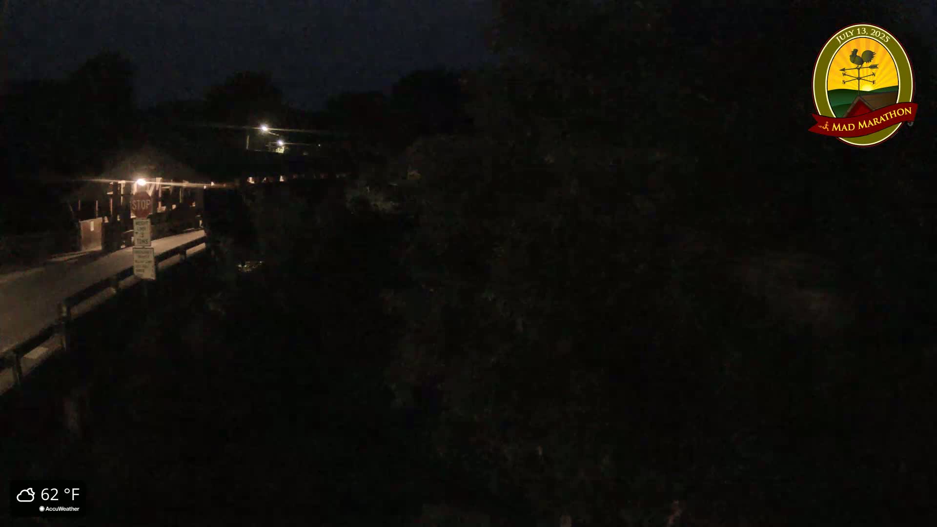 A nighttime, overhead view of a dark, tree-lined bridge with a stop sign visible, under a clear, 62°F sky.