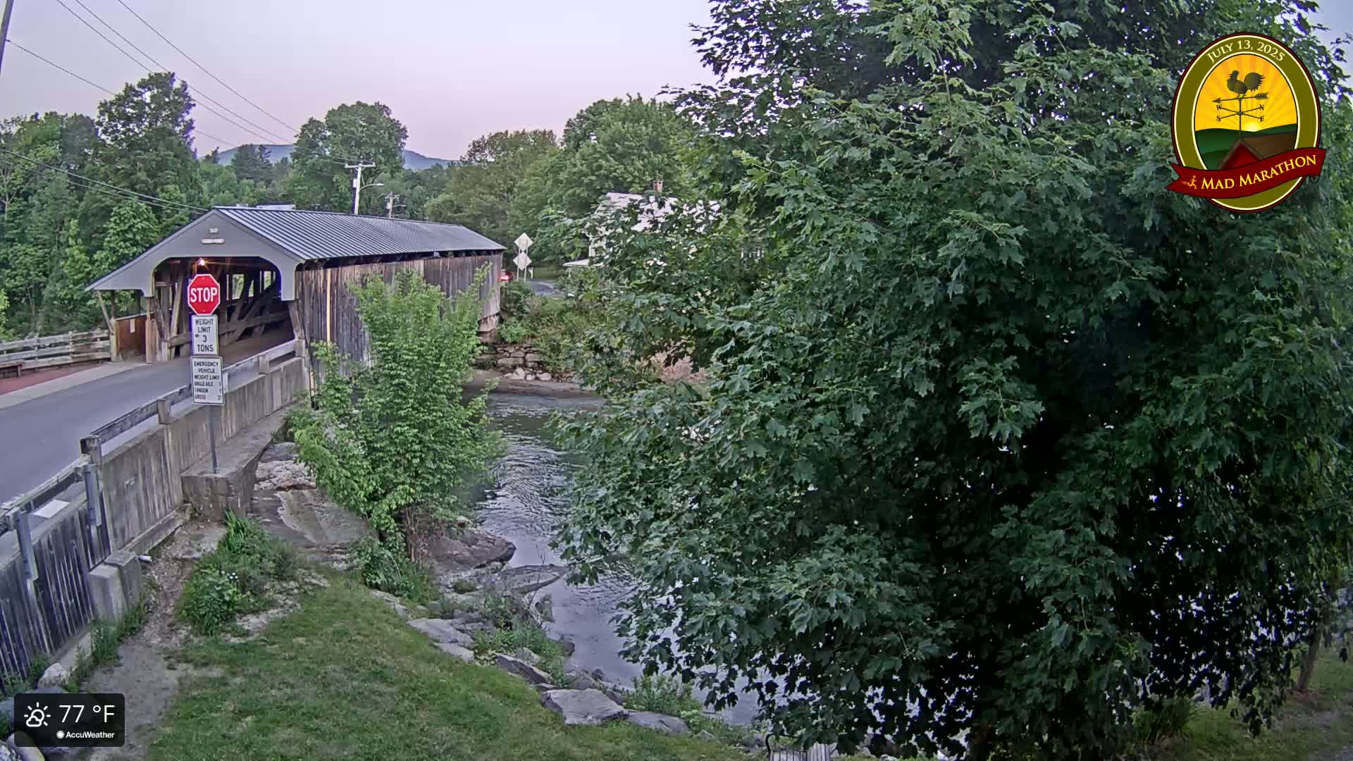 A covered bridge spans a calm river under a partly cloudy sky, surrounded by lush green trees and vegetation.